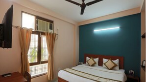 A hotel room with a bed, a ceiling fan, a TV, a window with curtains, and a bedside table with a lamp. - Hotel Glinzer Palace By Aura (Noida)