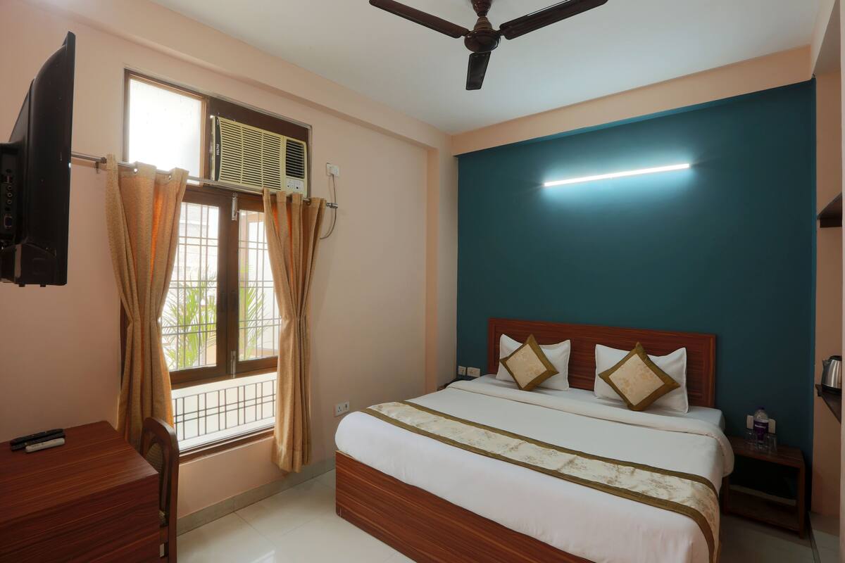 A hotel room with a bed, a ceiling fan, a TV, a window with curtains, and a bedside table with a lamp.