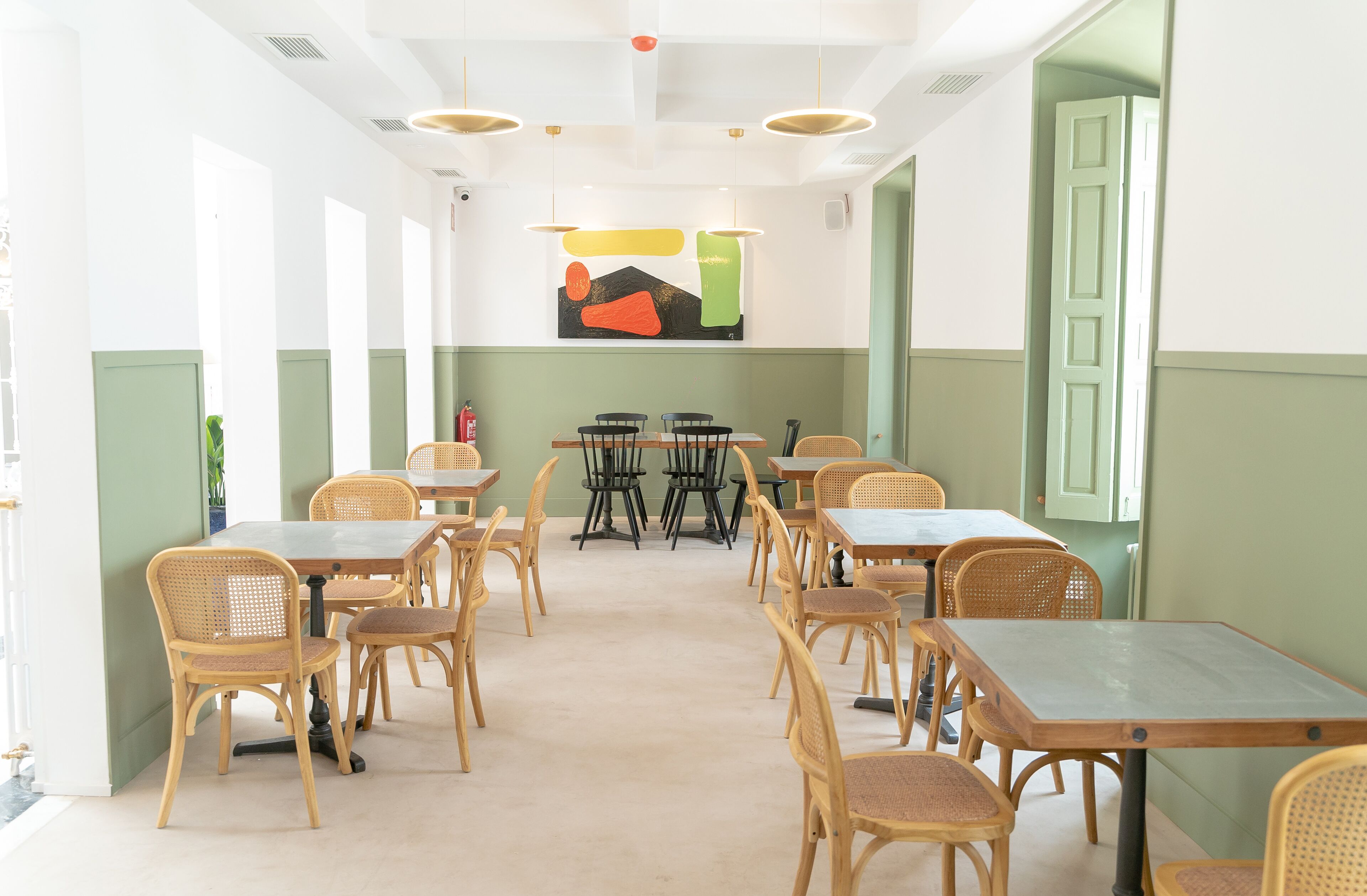 Een eetruimte met meerdere tafels en stoelen, een groot schilderij aan de muur, en een groene deur.