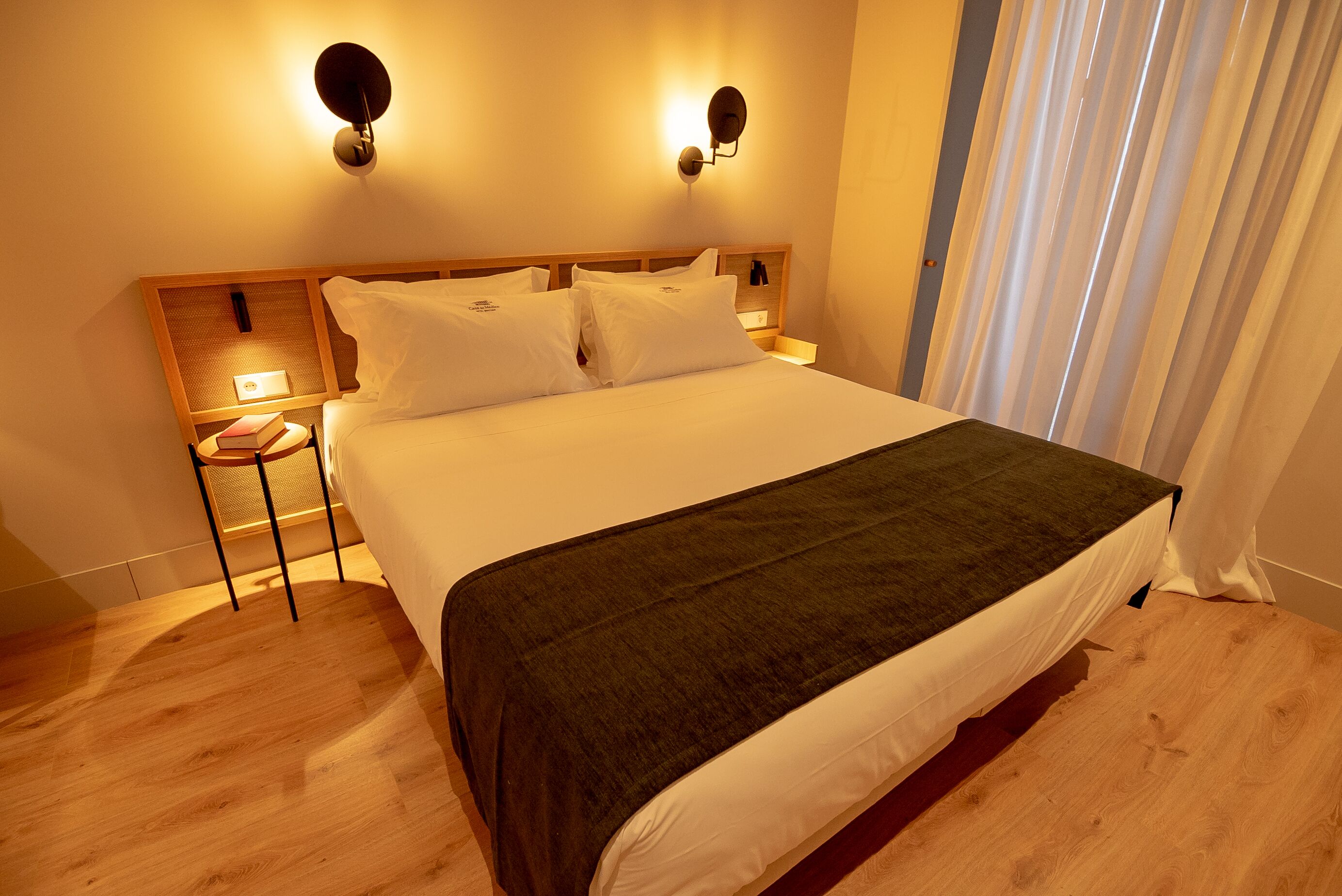 Una cama bien hecha con un edredón oscuro, dos lámparas fijadas a la pared y una mesita de noche con un libro y una lámpara.