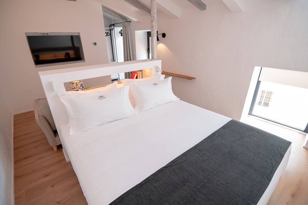 A modern bedroom with a large bed, a TV mounted on the wall, and a window providing natural light.