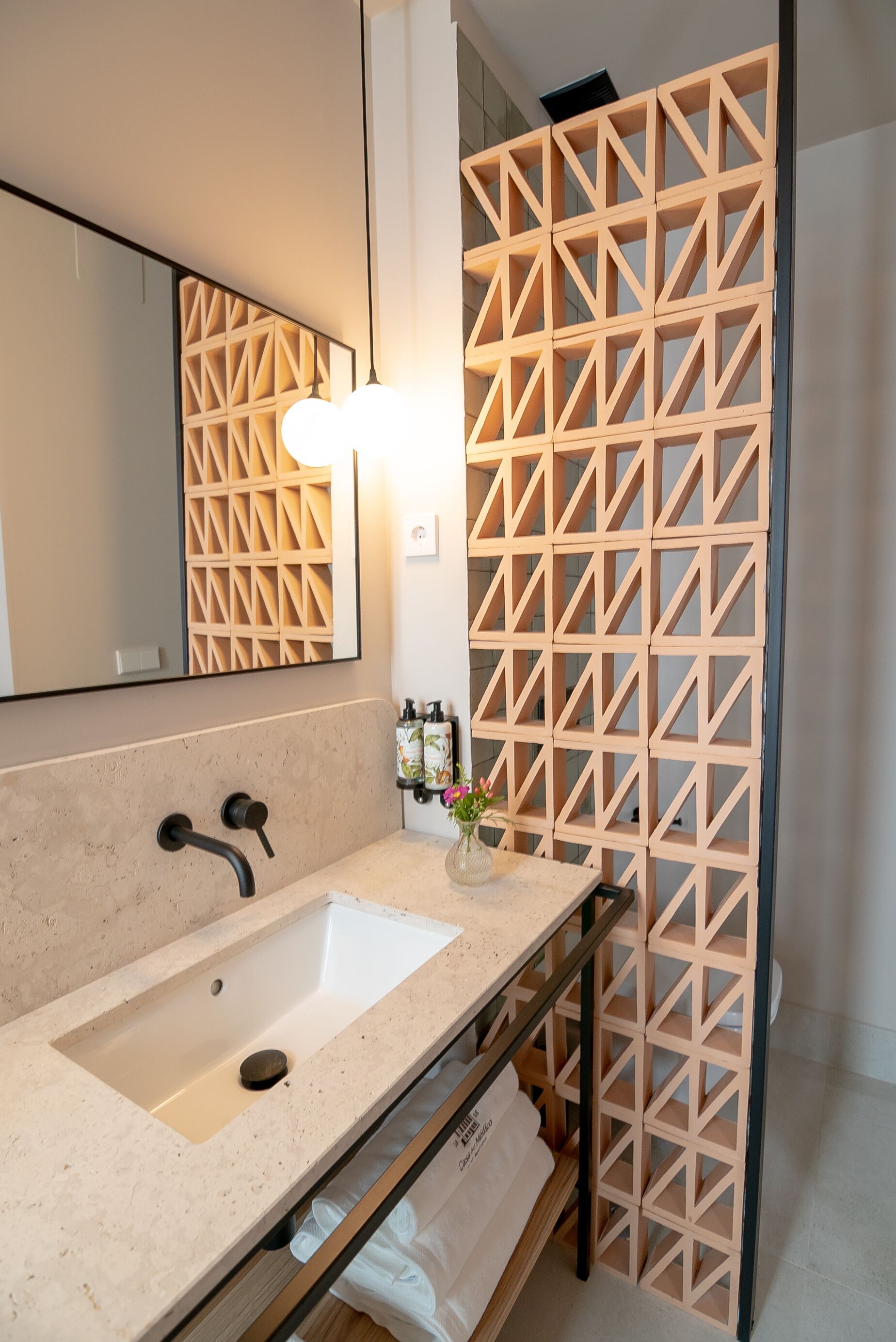 Un baño moderno con lavamanos de encimera color beige, un espejo y un separador de madera con un diseño geométrico.