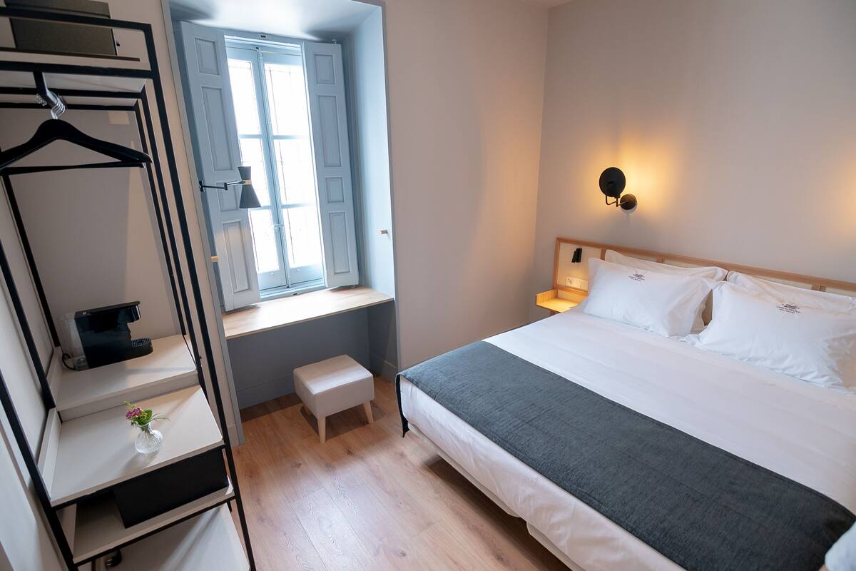 A bedroom with a bed, bedside table, lamp, window, and a hanging rack with a small vase.