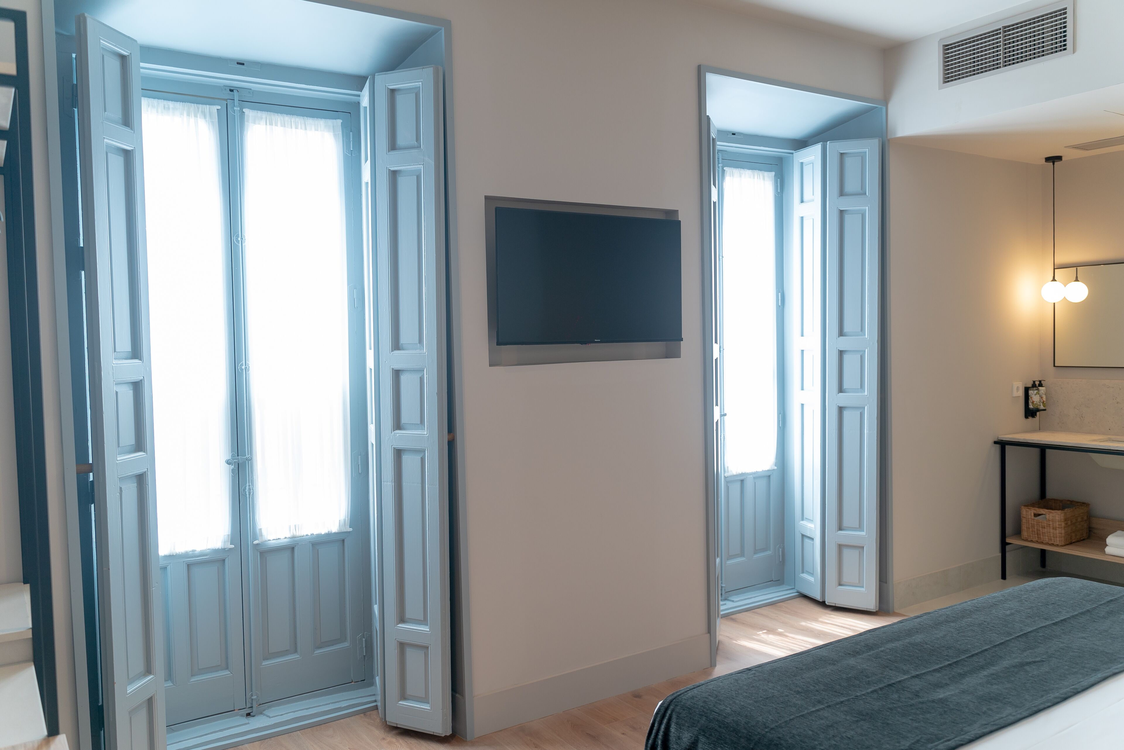 A room with a large window, a flat-screen TV mounted on the wall, and a bedside table with a lamp.
