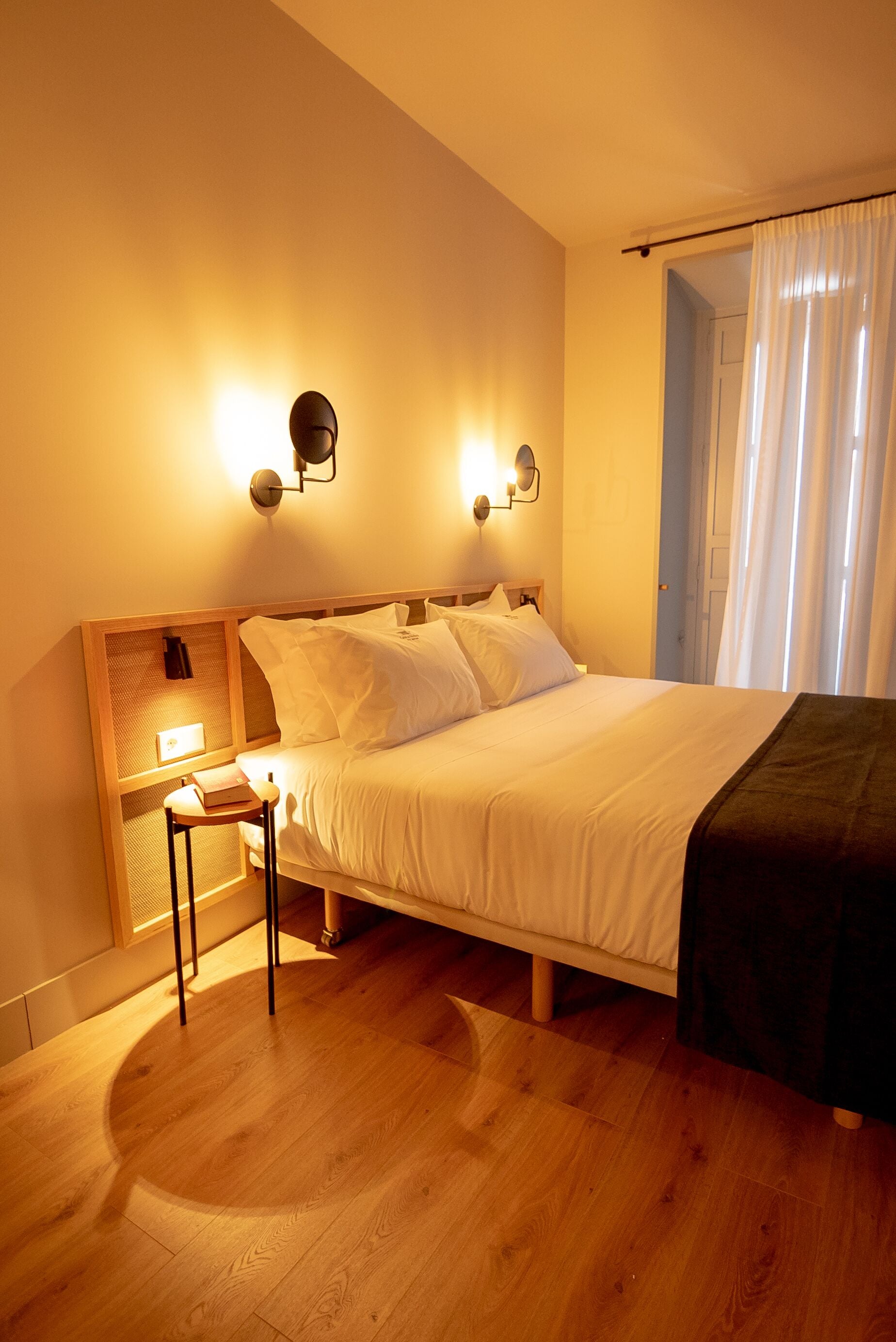 Ein ordentlich eingerichtetes Schlafzimmer mit einem hölzernen Kopfende, einem Nachttisch mit Lampe und einem Fenster mit Vorhängen.