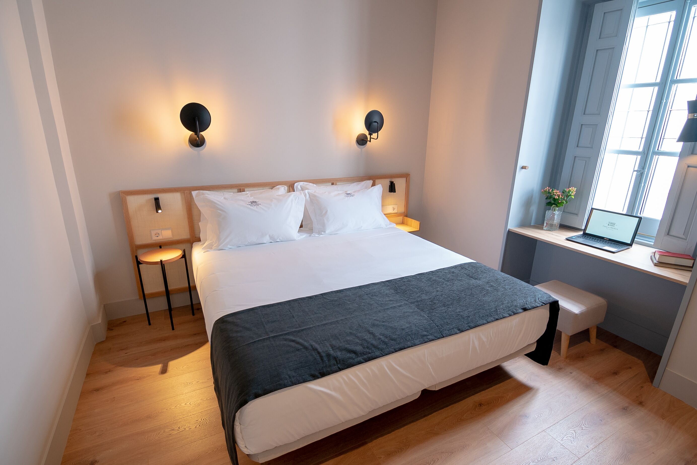 Een netjes ingerichte slaapkamer met een groot bed, nachtkastjes, een bureau met een laptop en een raam met natuurlijk licht.