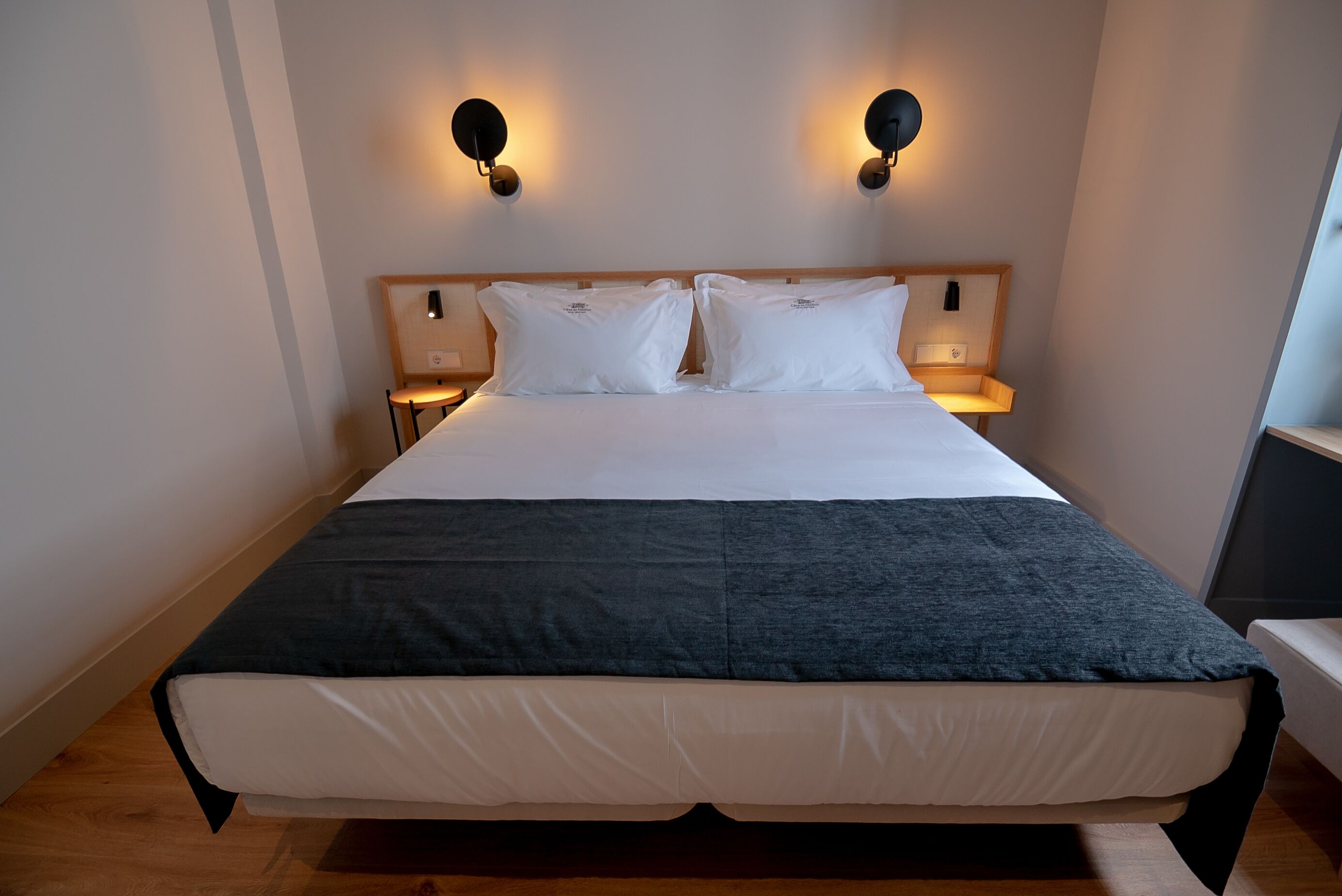 Una cama bien tendida con ropa de cama blanca y gris oscuro, dos mesitas de noche fijadas a la pared y un cabecero de madera.