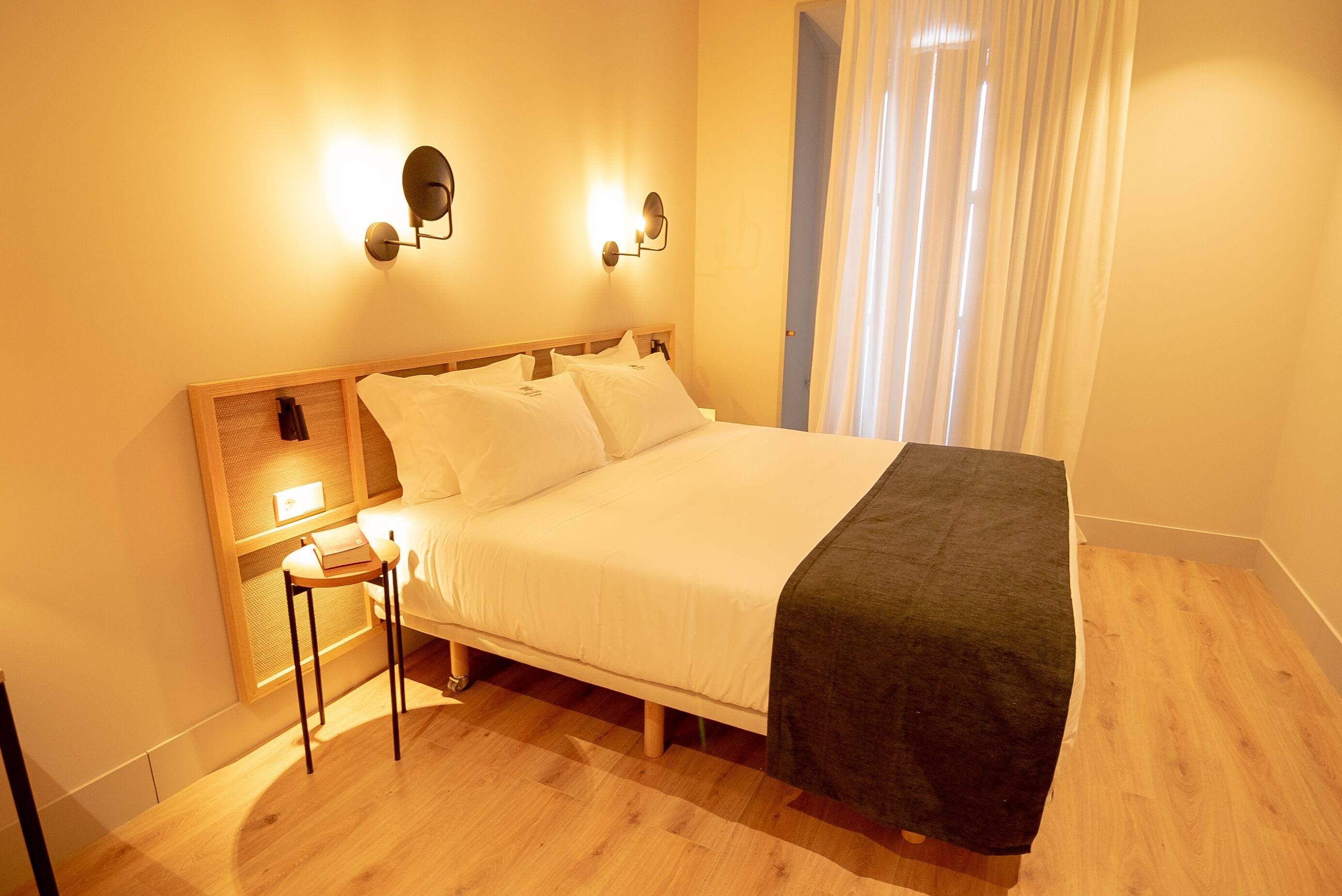 Una cama bien hecha con sábanas blancas, una mesita de noche con una lámpara y una ventana con cortinas de gasa.