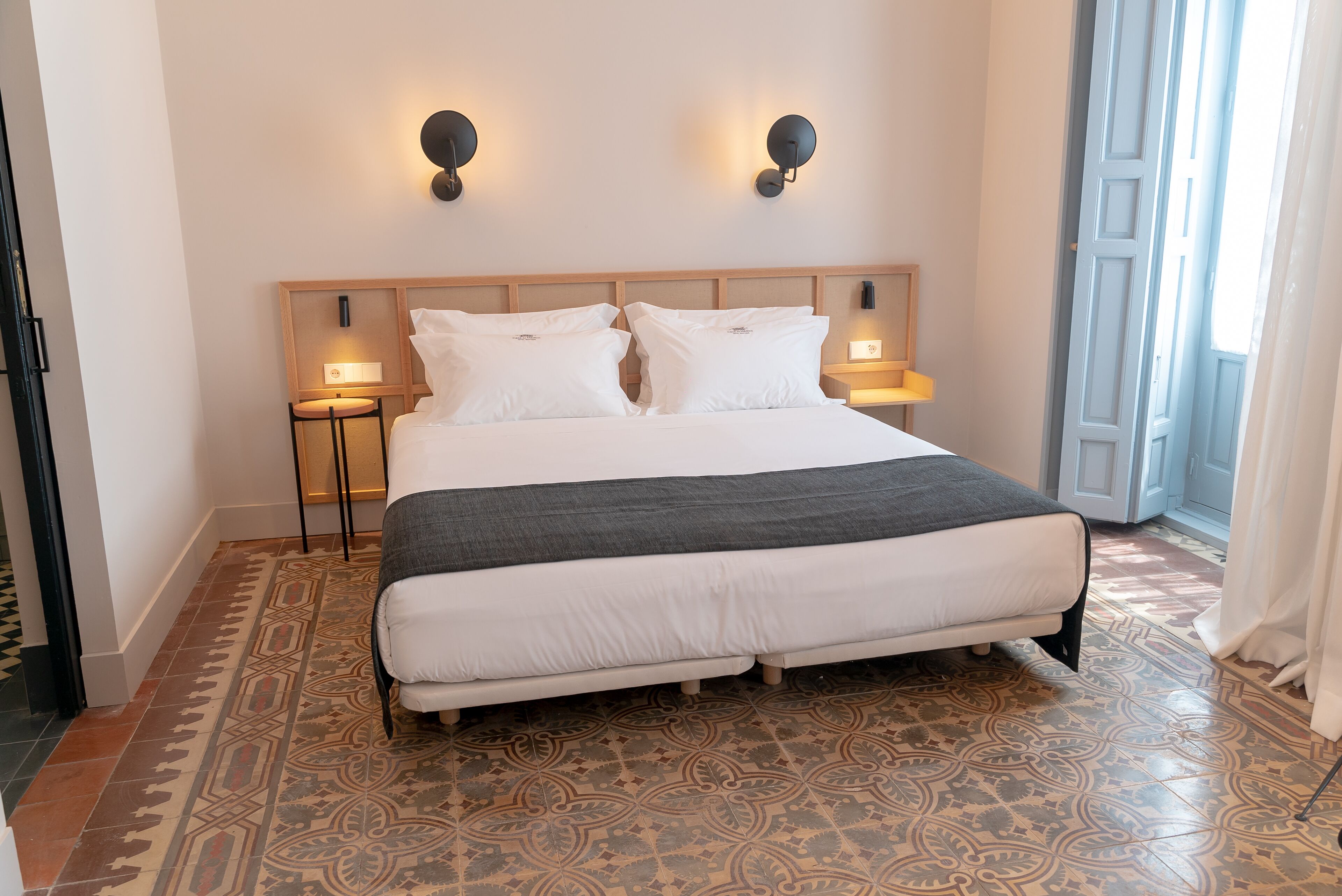 A hotel room with a large bed, two bedside tables with lamps, patterned floor tiles, and a wooden headboard.
