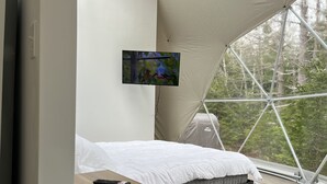 Une chambre aménagée comme une tente, avec un lit, un téléviseur fixé au mur et une vue sur la forêt.