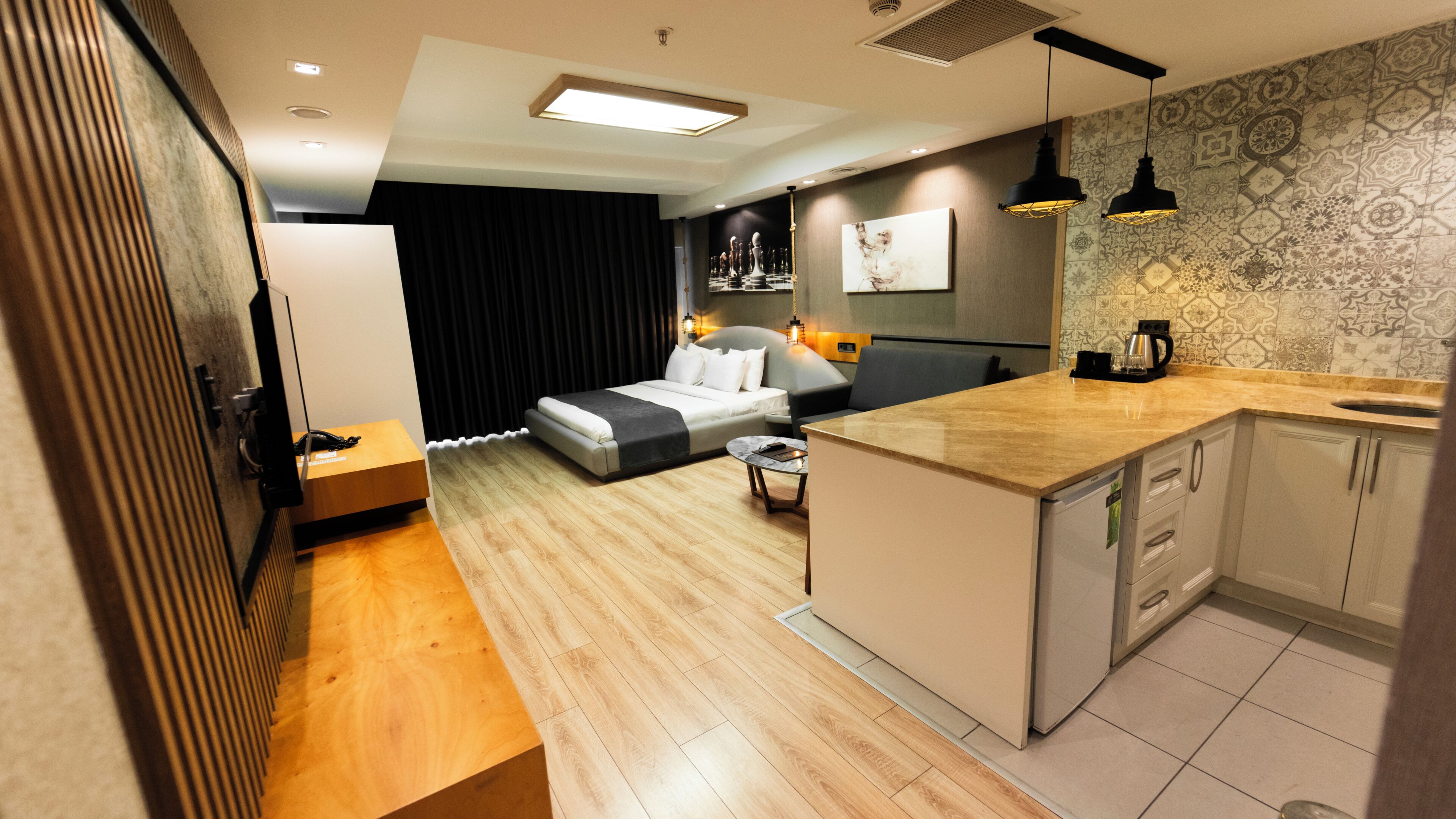 A modern hotel room with a bed, a small dining table, a kitchen area, and a living area with a sofa.