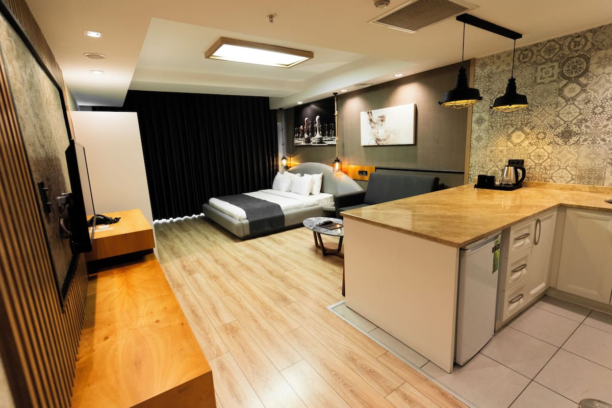 A modern hotel room with a bed, a small dining table, a kitchen area, and a living area with a sofa.