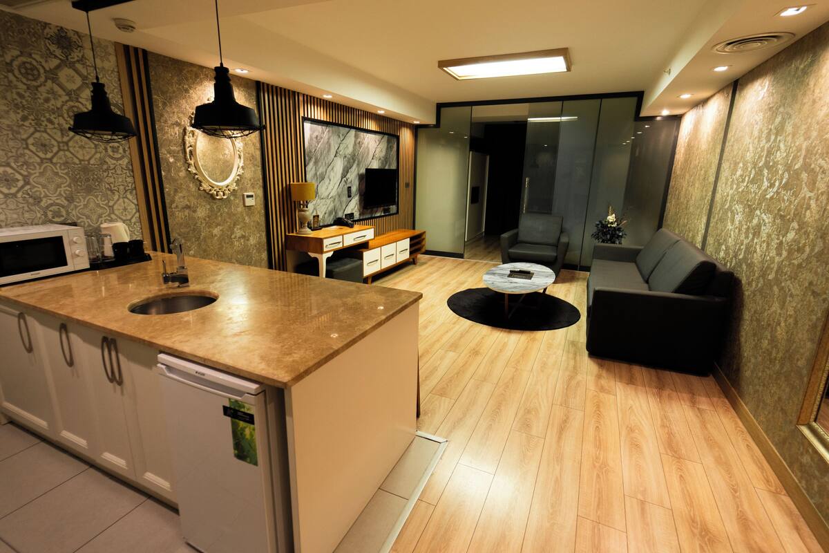 A modern kitchen with a built-in microwave, a sink, and a wooden countertop. There is a dining area with a round table and a sofa. The room has patterned wallpaper and a large mirror.