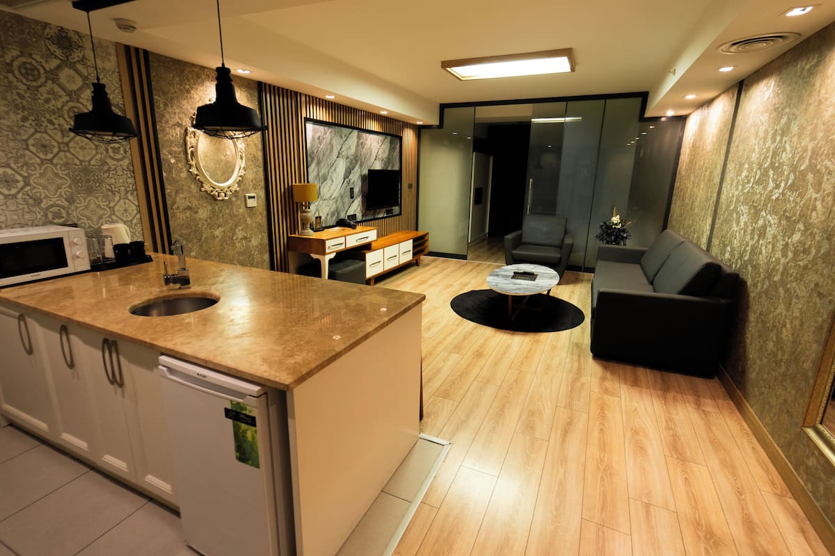 Una cocina moderna con microondas integrado, un fregadero y una encimera de madera. Hay una zona de comedor con una mesa redonda y un sofá. La habitación tiene papel tapiz estampado y un espejo grande.