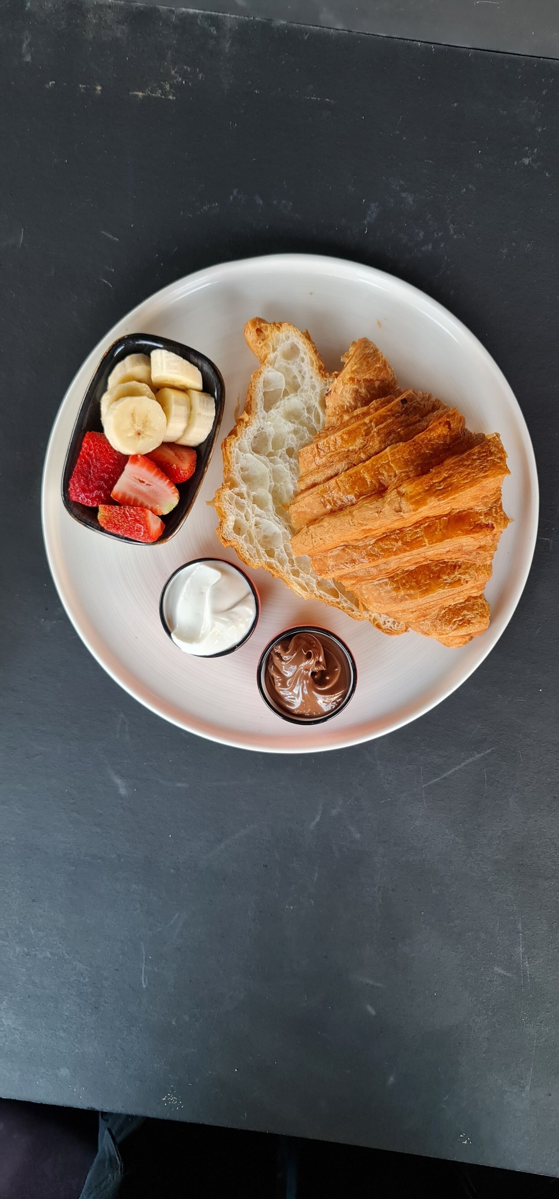 A plate with croissant, sliced bananas, strawberries, and two types of spreads on a dark surface.