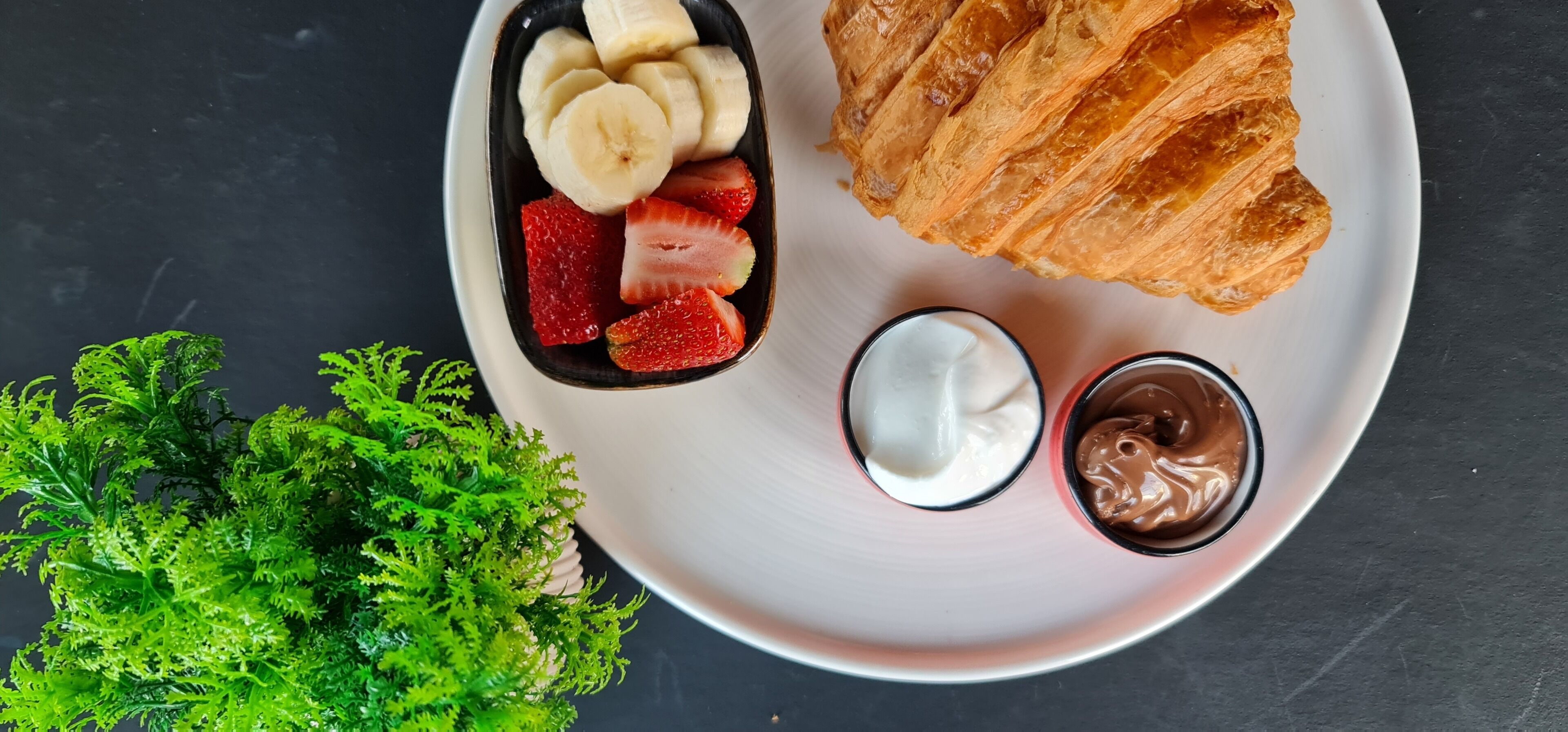 A plate with a croissant, sliced bananas, strawberries, and two small bowls of cream and chocolate sauce.