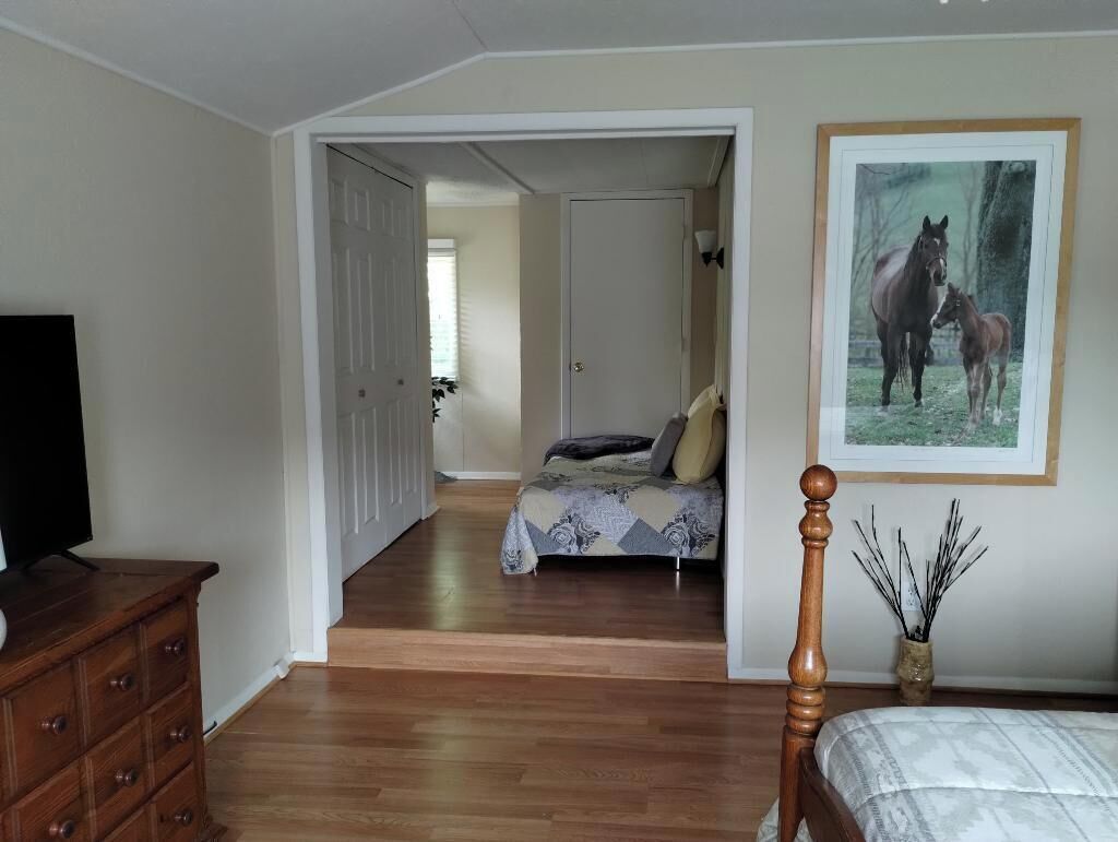 Une chambre avec une commode en bois, un lit recouvert d’une couvre-lit à motifs, un tableau encadré représentant des chevaux et un couloir menant à une autre pièce.