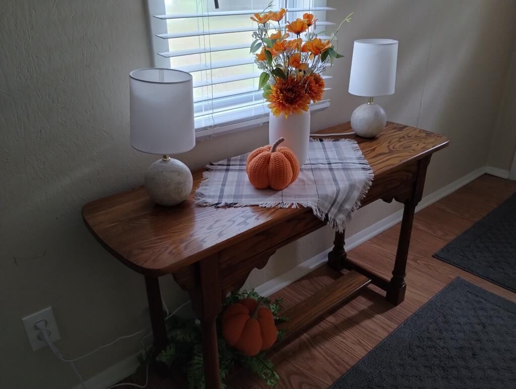Ett konsolbord i trä med en vas med orange blommor, två lampor och en liten prydnad i form av en pumpa.