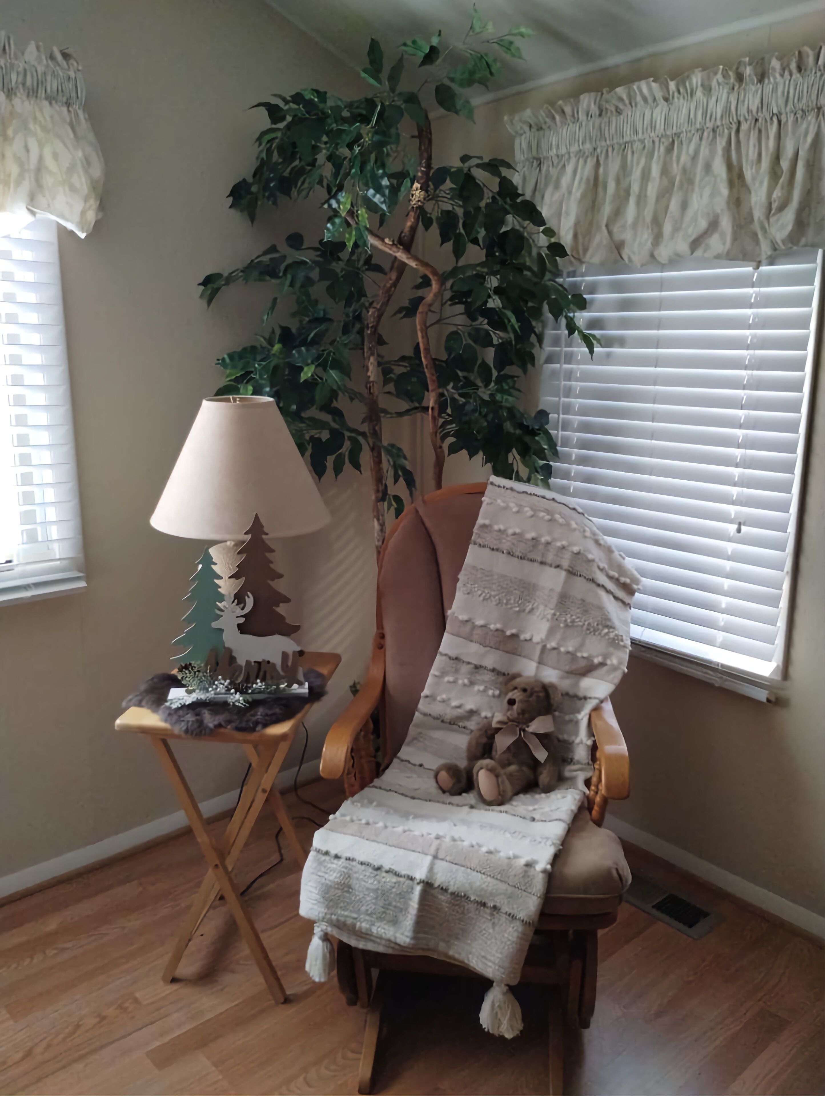 Ein gemütliches Zimmer mit einem hölzernen Stuhl, der mit einer weißen Decke bedeckt ist, einem kleinen Tisch mit einer Lampe, einem dekorativen Baum und einer großen Topfpflanze.