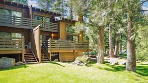 A modern log cabin with a wooden deck, a staircase leading to a second level, and a surrounding forest. - Rocky Ridge Condo: Walk to Lake, Pools & Trails (Tahoe City)