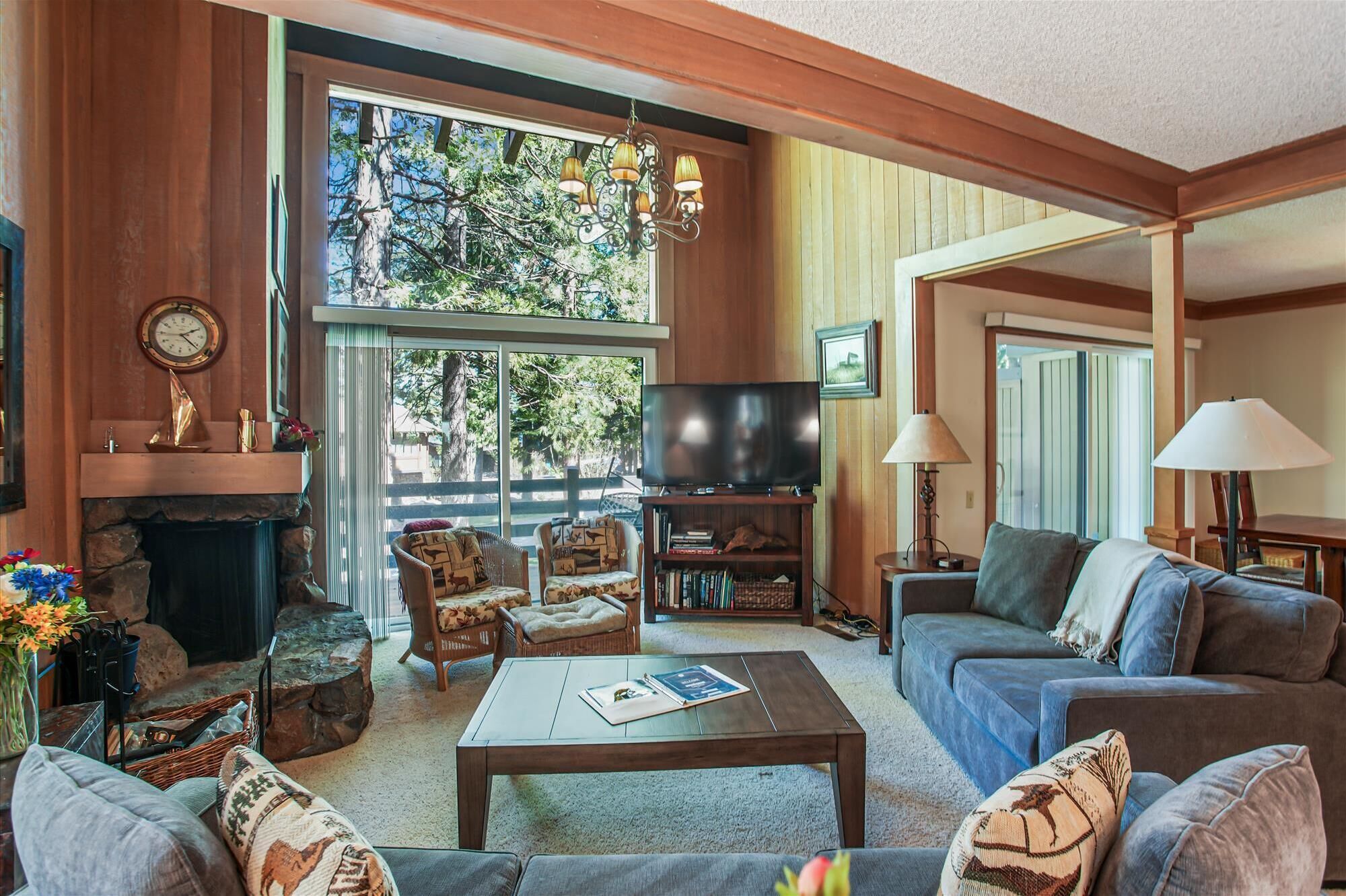 A cozy living room with a fireplace, comfortable seating, a television, and a large window offering a view of trees.