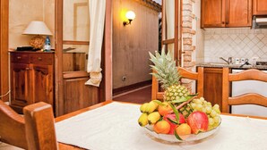 A dining area with a table set for two, a fruit bowl, wooden chairs, a lamp, and a kitchen in the background. - Stunning Views of Baiadebahas Residenza-t6 2 Bedroom Apartment Sleeps 6 Persons (Golfo Aranci)
