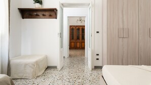 A hallway with a bed on the left, a wooden wardrobe on the right, and a cabinet with a vase of flowers above it.
