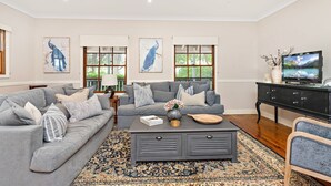A living room with a grey sofa set, a coffee table with decorative items, a TV on a black stand, and artwork on the walls. - Anembo Hideaway - Relaxing Oasis (Kiama Heights)