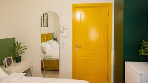 A hotel room with a bed, a mirror, a yellow door, a white bedside table with a plant, and a green wall. - Room-three Doors Apartments, Papaya 1-be (Tirana)