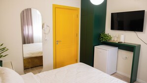 A hotel room with a bed, a yellow door, a small fridge, a TV, and a plant. - Room-three Doors Apartments, Papaya 1-be (Tirana)