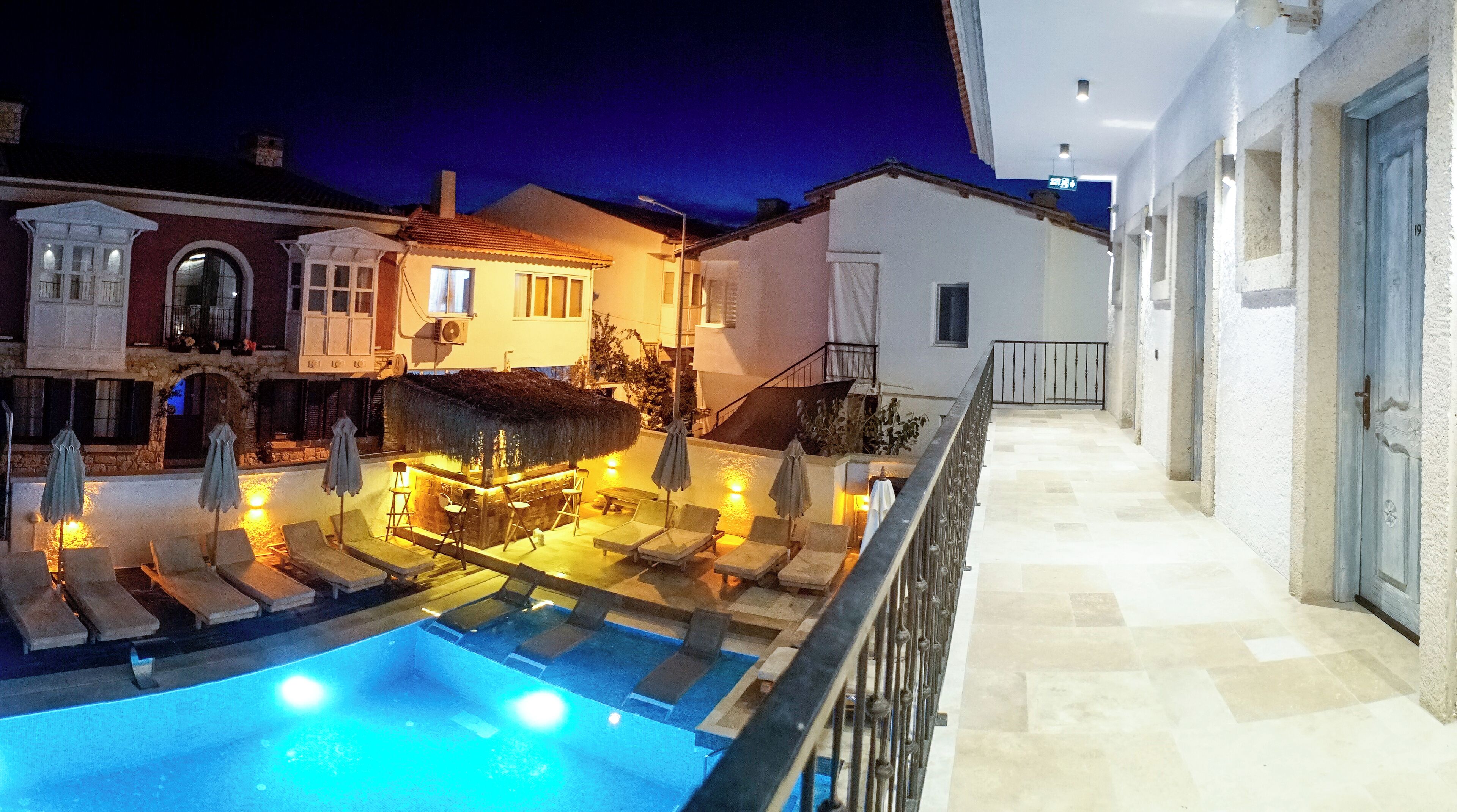 A pool area with lounge chairs and a bar, surrounded by buildings with balconies and a staircase leading to an upper level.