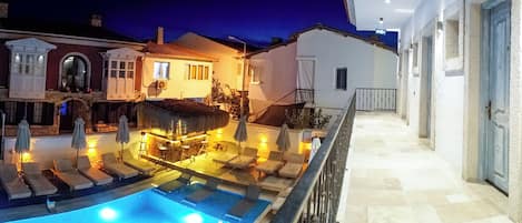 A pool area with lounge chairs and a bar, surrounded by buildings with balconies and a staircase leading to an upper level.