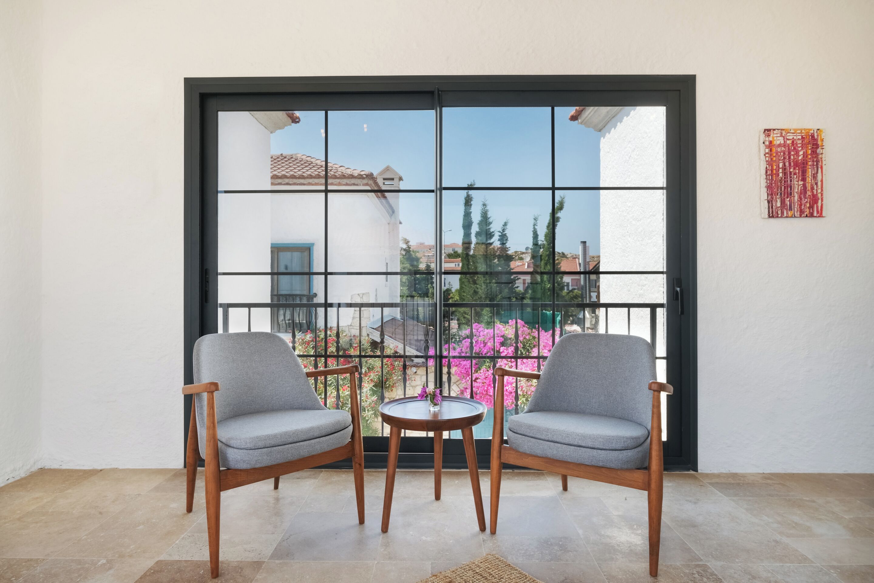 A balcony with two grey chairs, a small round table, and a view of a garden with pink flowers.