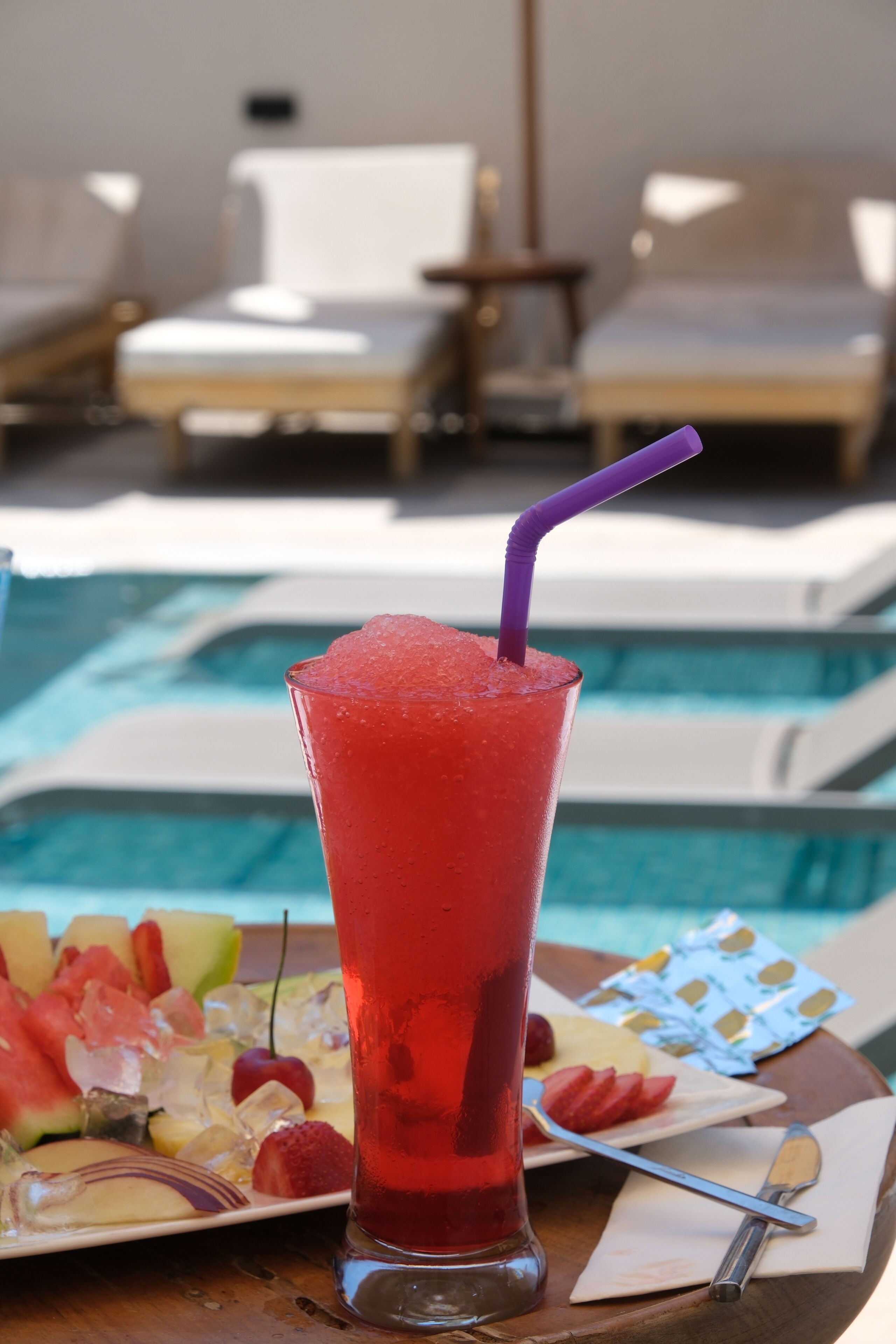 A poolside table with a tall glass of red beverage, fruit, and a deck of playing cards.