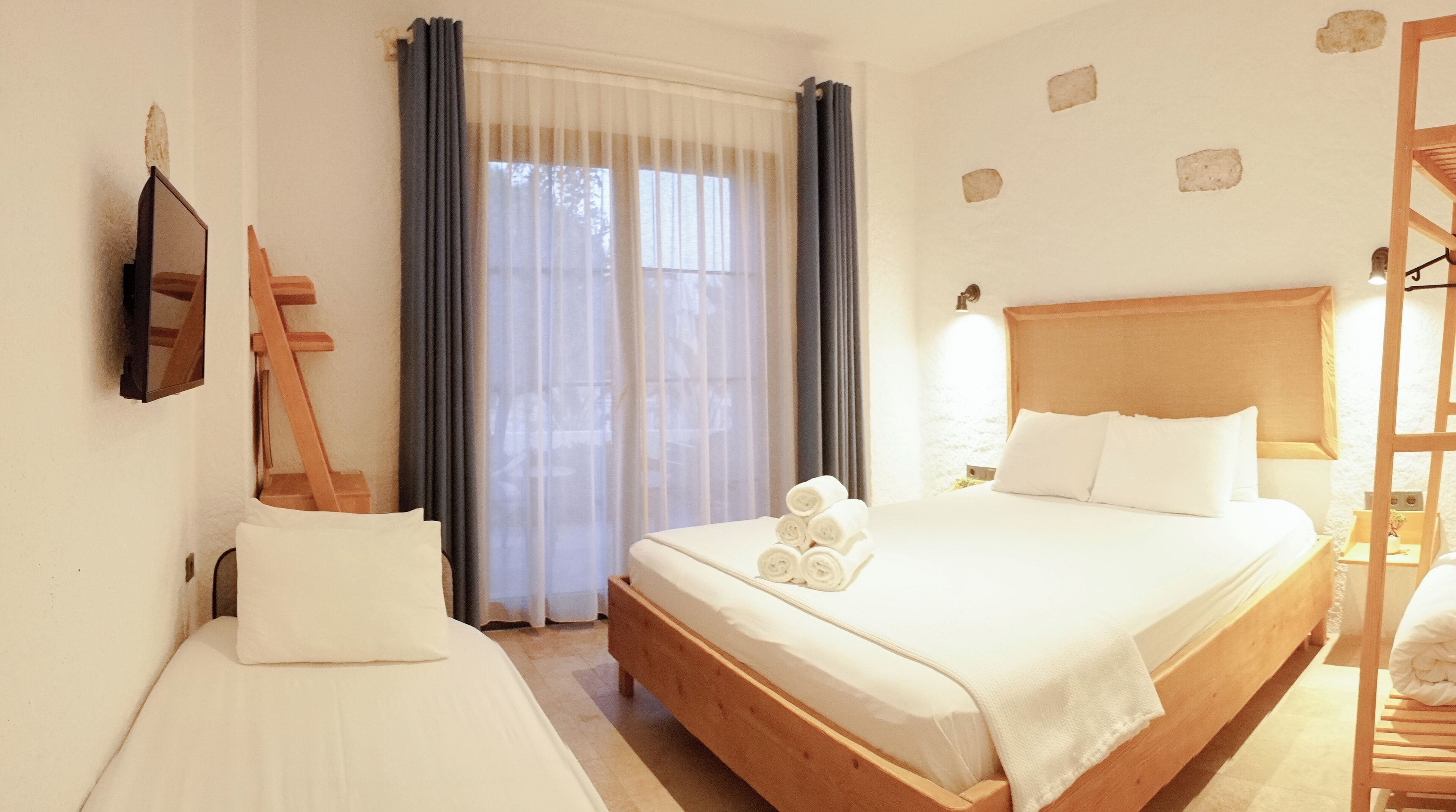 A hotel room with a bed, a smaller bed or sofa, a wooden ladder, a wall-mounted TV, and a window with curtains.