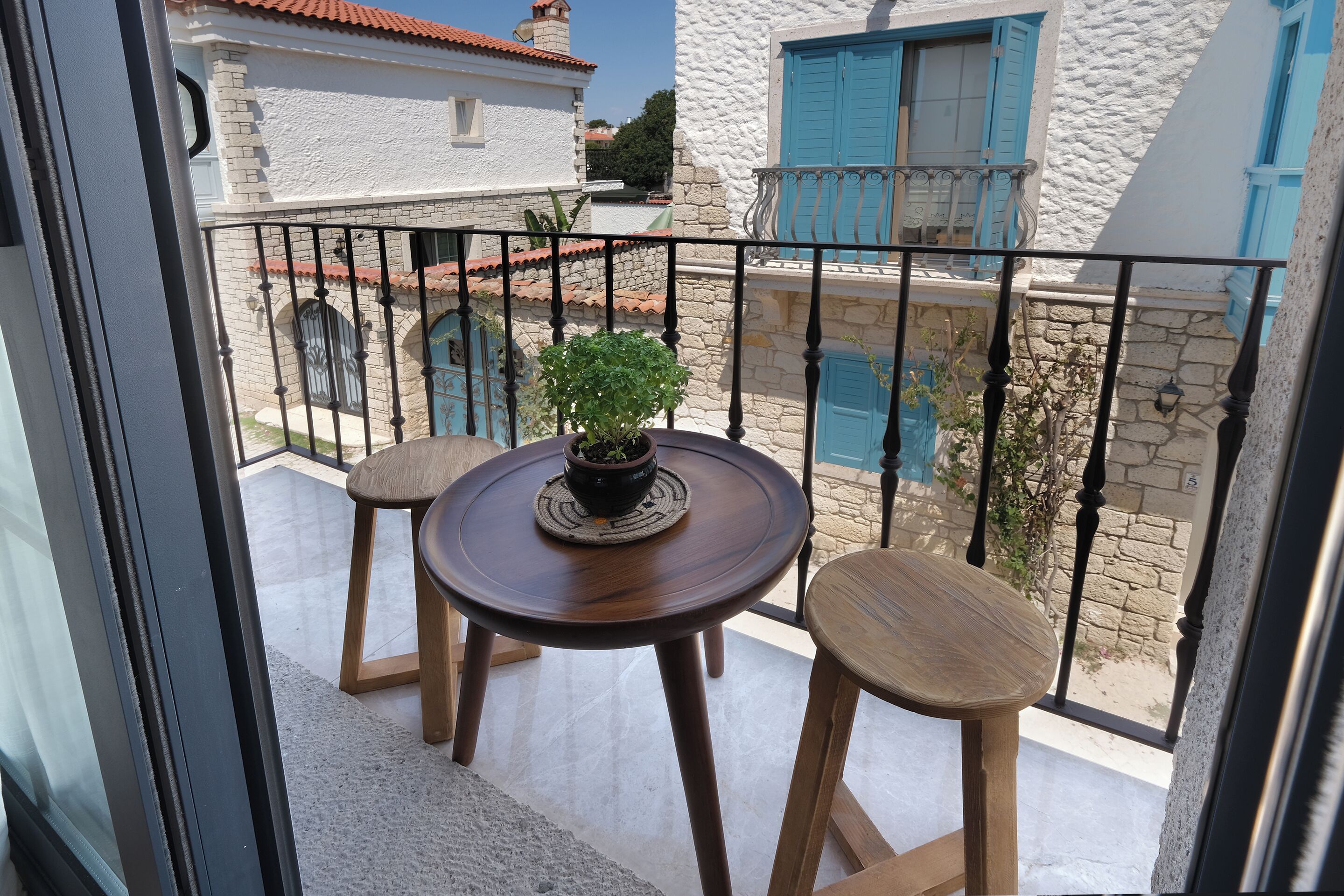 A balcony with a round wooden table and two wooden stools, a potted plant, and a view of white buildings with blue shutters.