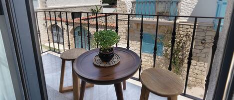 A balcony with a round wooden table and two wooden stools, a potted plant, and a view of white buildings with blue shutters.