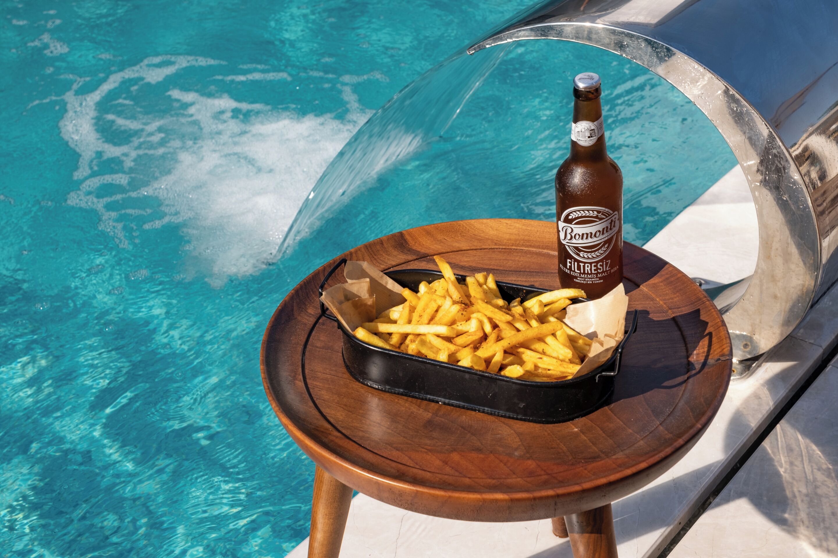 A wooden tray with a bottle of beer and fries by a poolside.