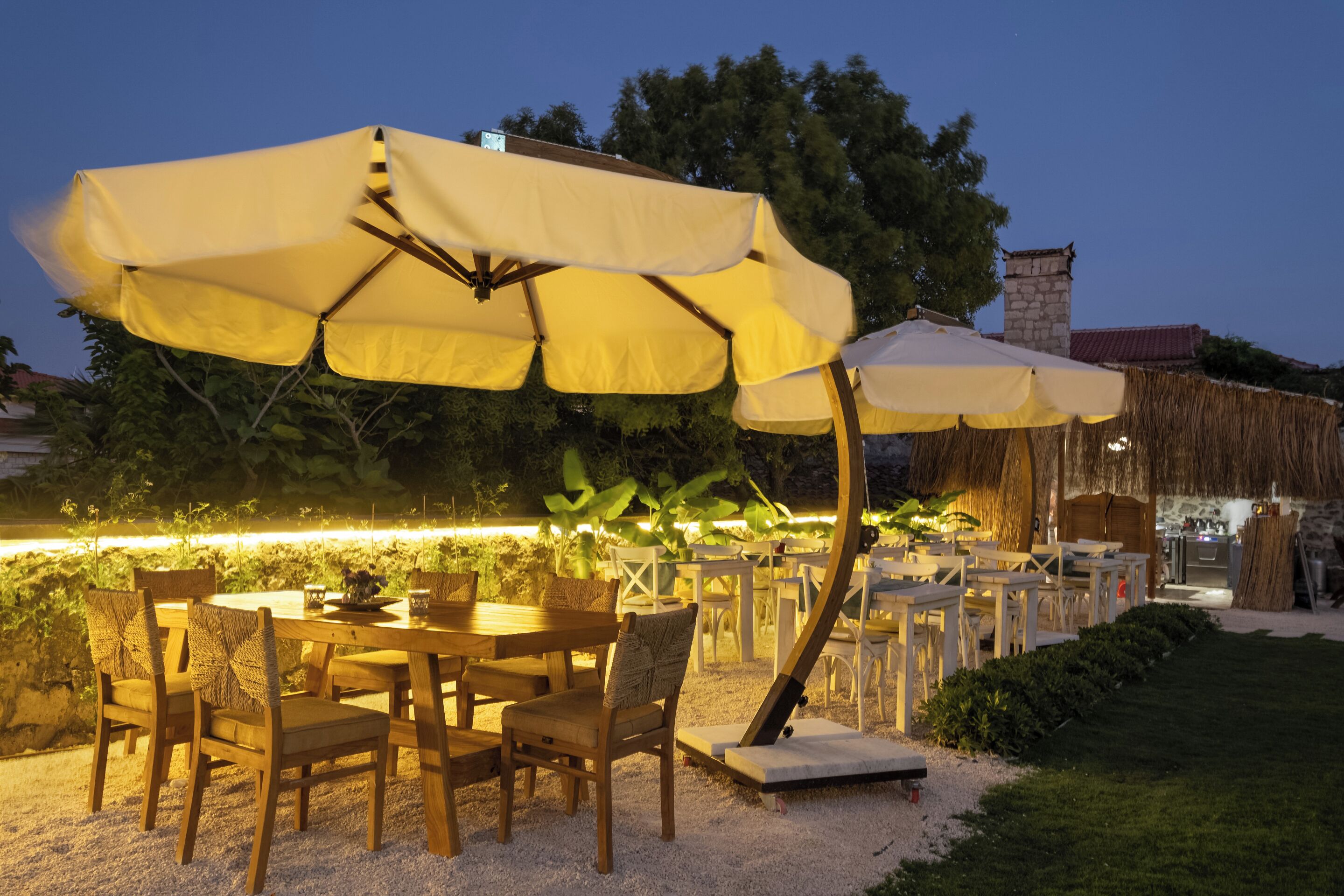 Un área de comedor al aire libre con mesas y sillas de madera, cubierta por grandes sombrillas amarillas, y un edificio de piedra al fondo.