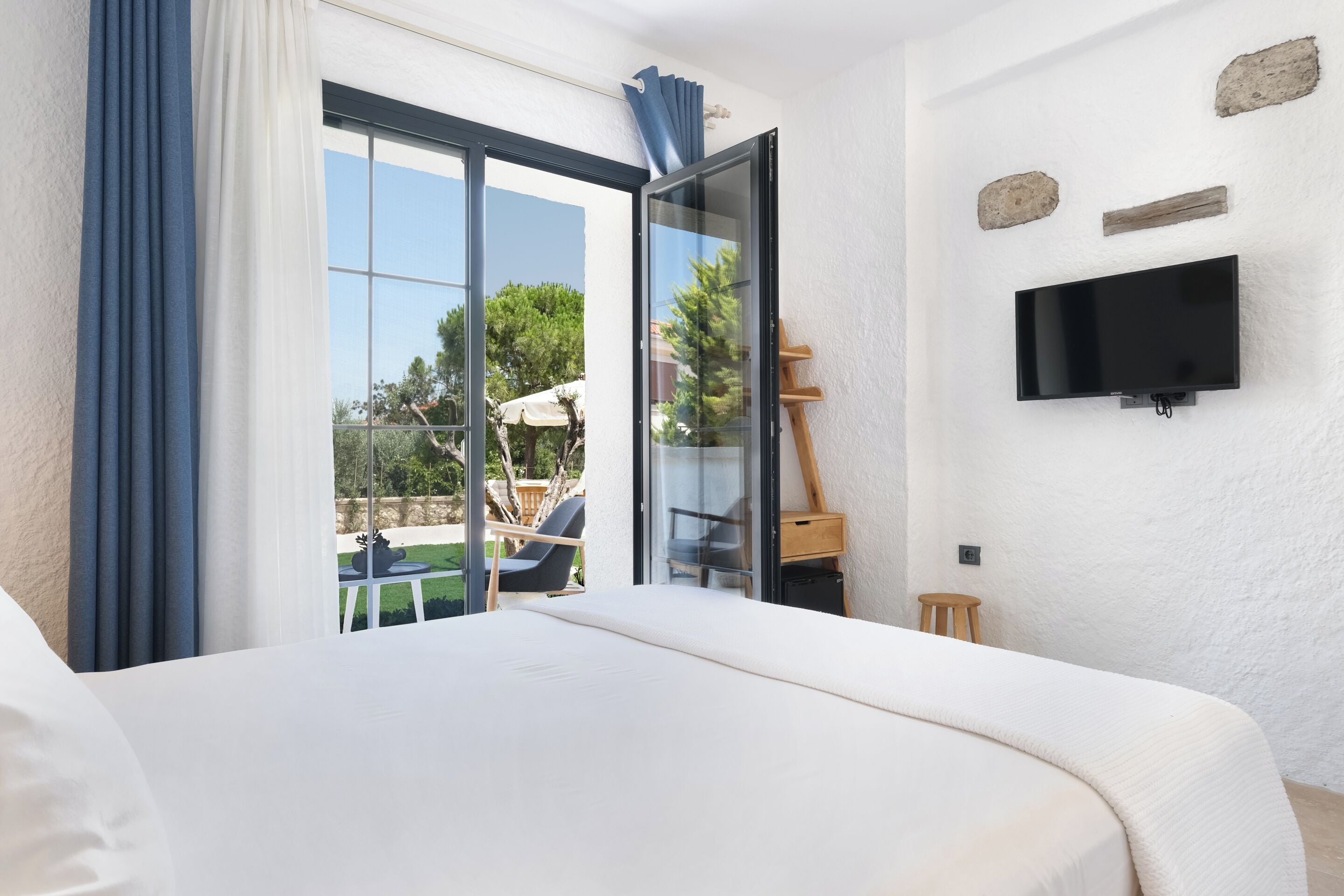 A bedroom with a large bed, a sliding glass door to a balcony, a flat-screen TV mounted on the wall, and a small wooden stool.