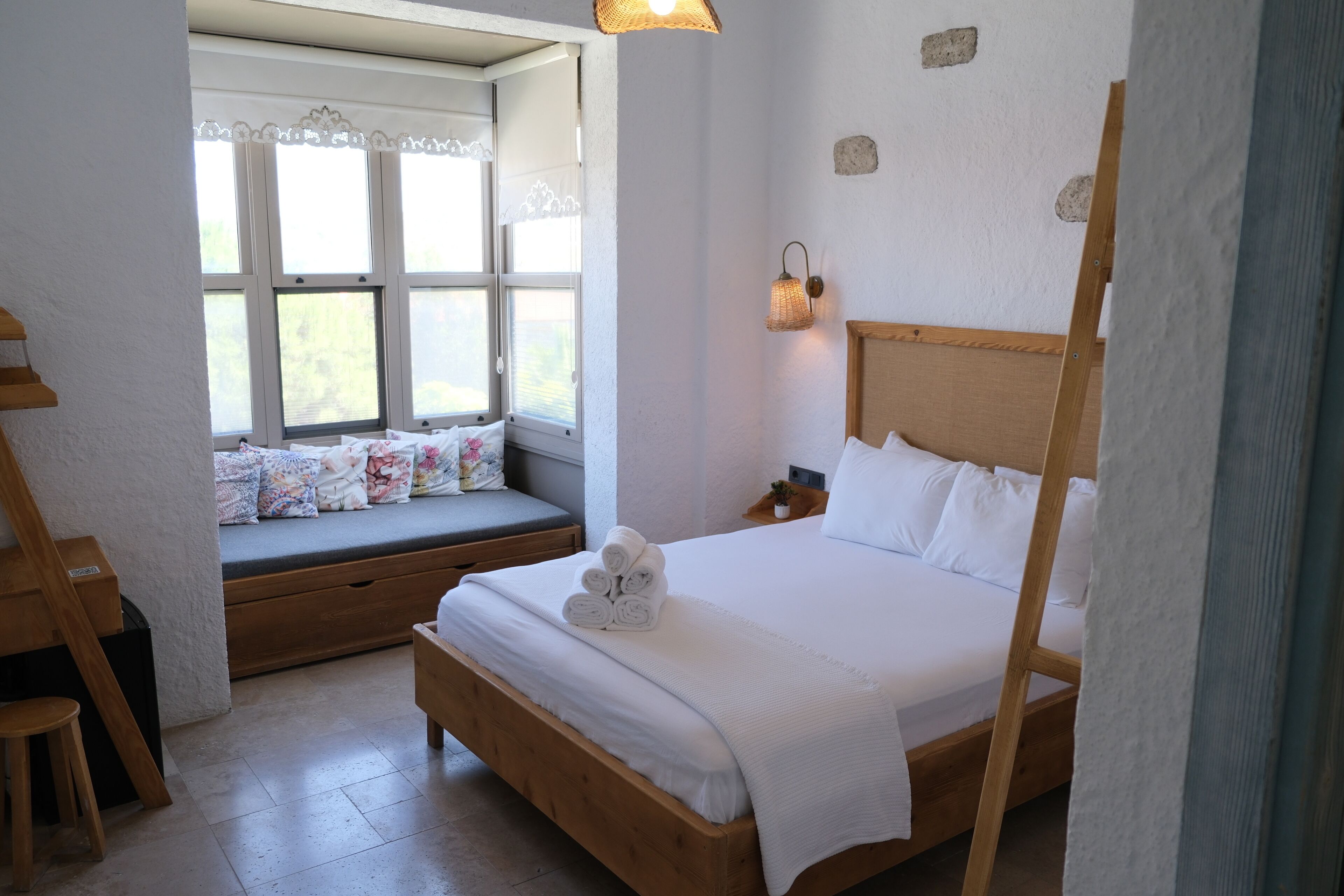 Un dormitorio con cama de madera, un rincón con ventanilla y cojines, una mesita de noche y una mesita auxiliar.