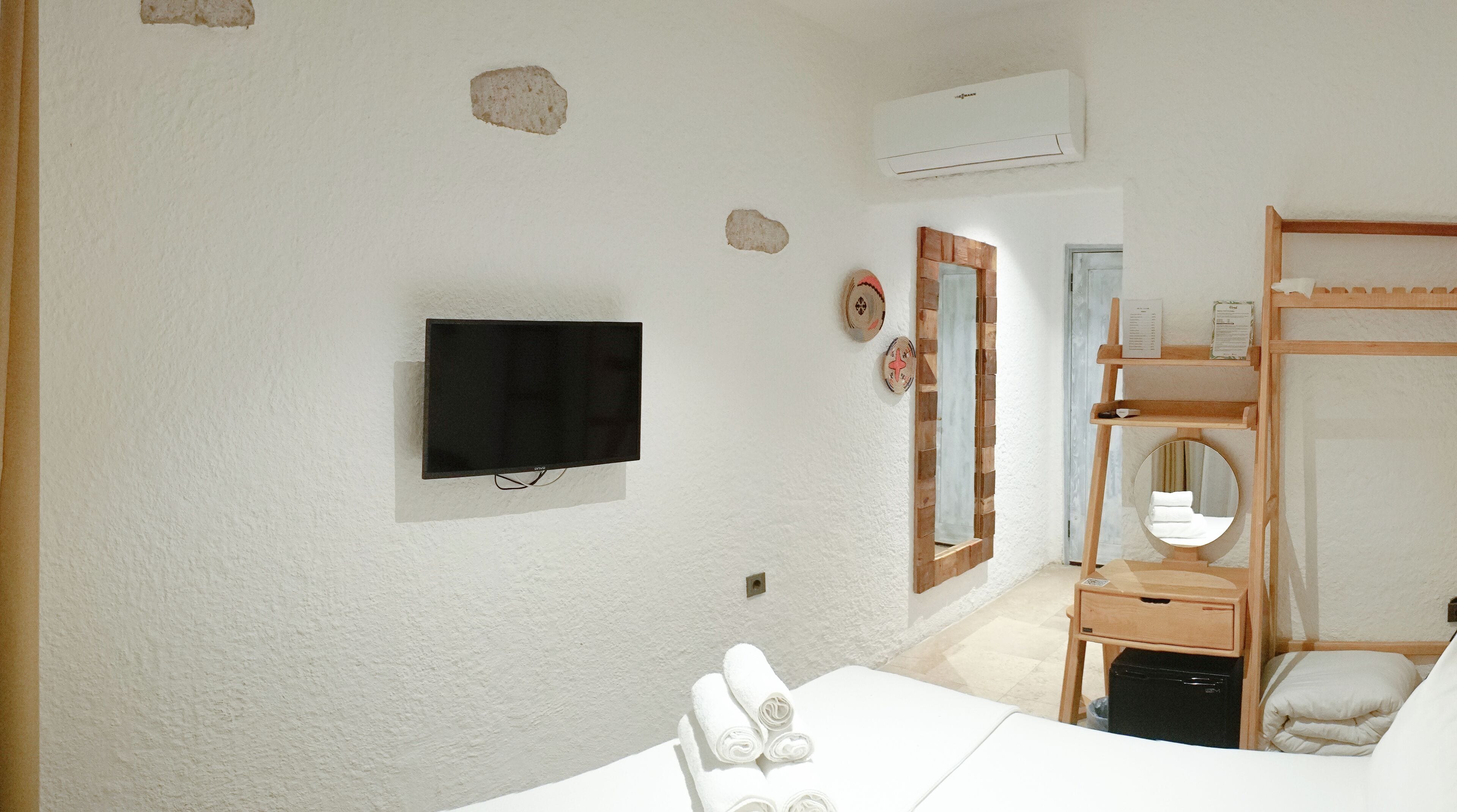 Une chambre d’hôtel équipée d’une télévision à écran plat, d’une étagère en bois où sont posés les articles de toilette, d’un miroir et d’un lit avec du linge de lit blanc.