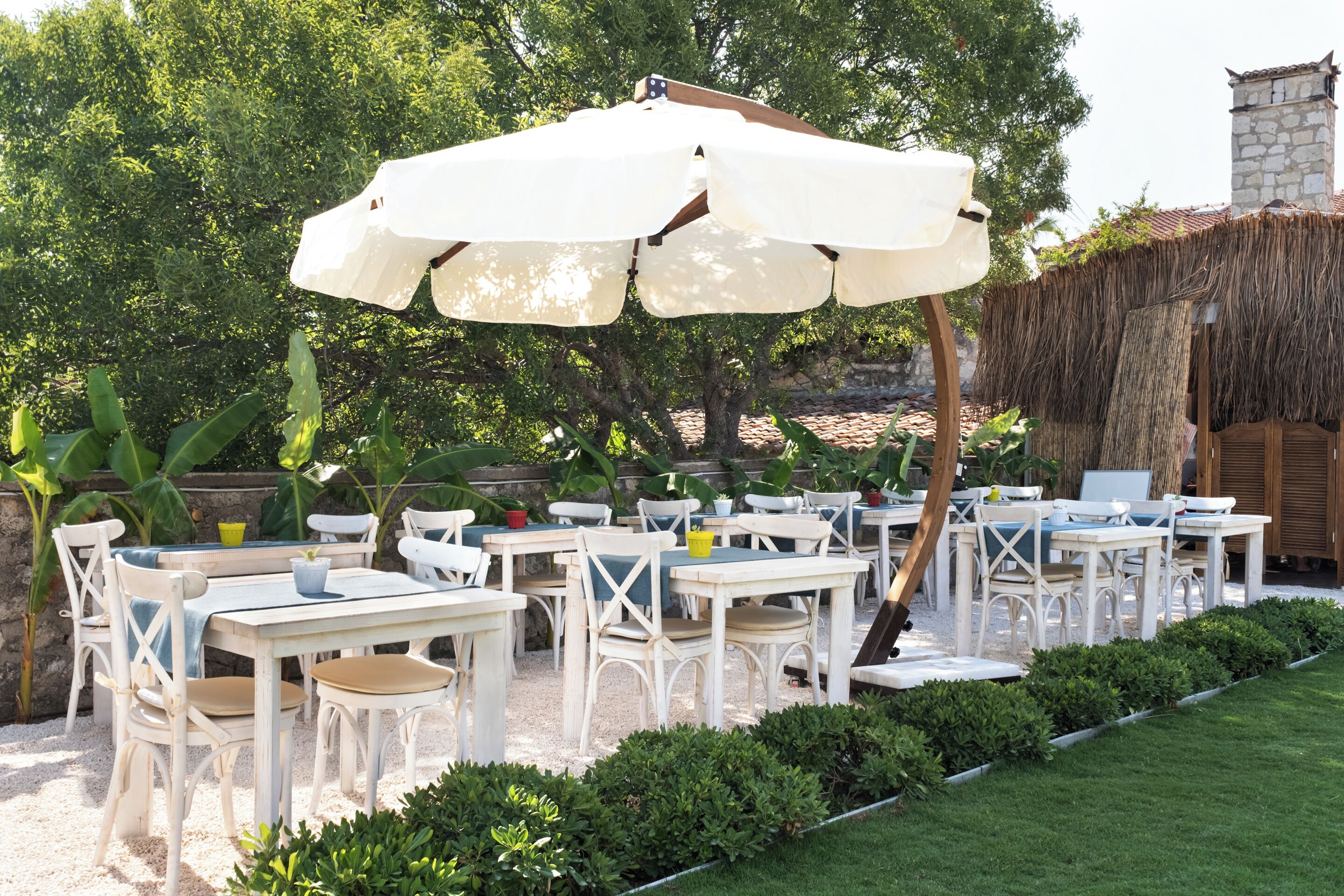An outdoor seating area with white tables and chairs, a large umbrella, and a thatched-roof structure in the background.