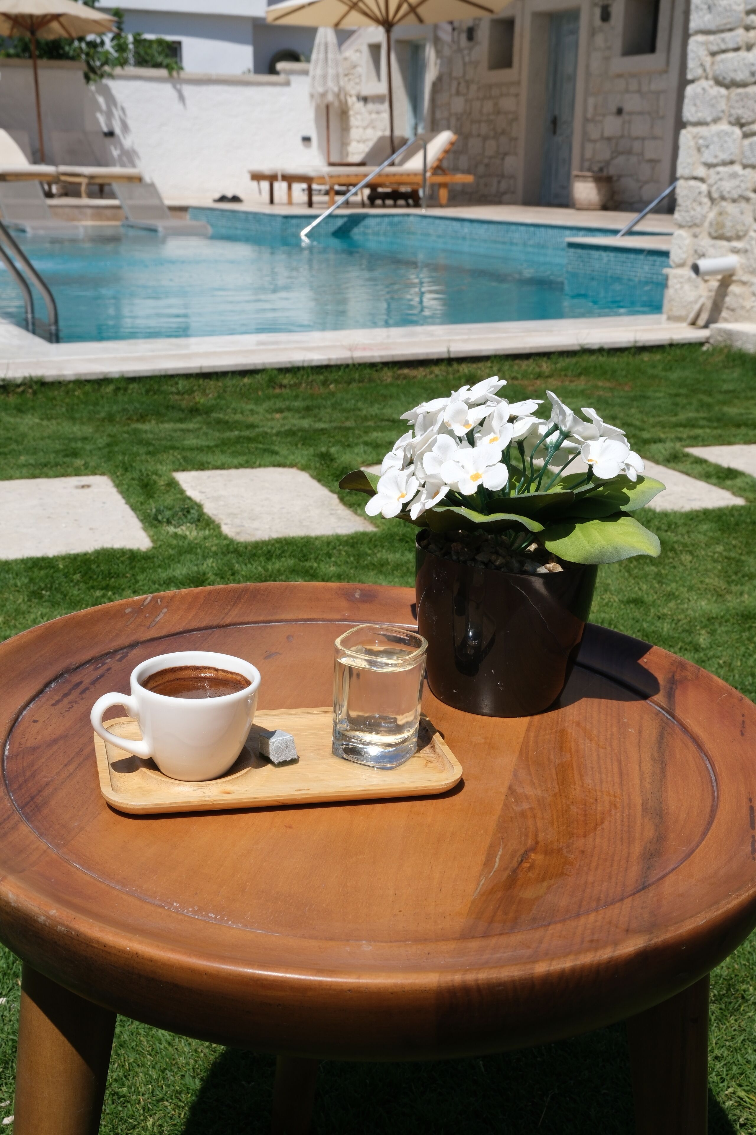 A wooden table with a white cup and a glass of water, a potted plant with white flowers, and a pool in the background.