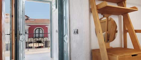 A hallway with open doors leading to a balcony, a wooden ladder, and a view of a building with a red roof.