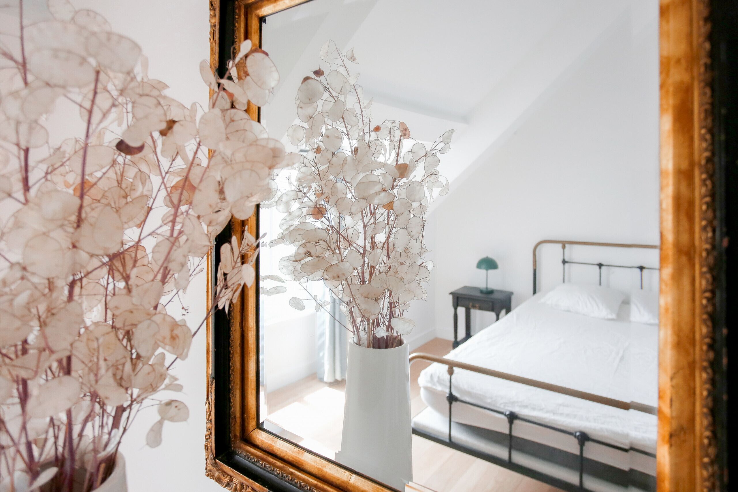 Ett sovrum med en säng, ett sängbord och en spegel som reflekterar en vas med torkade blommor.