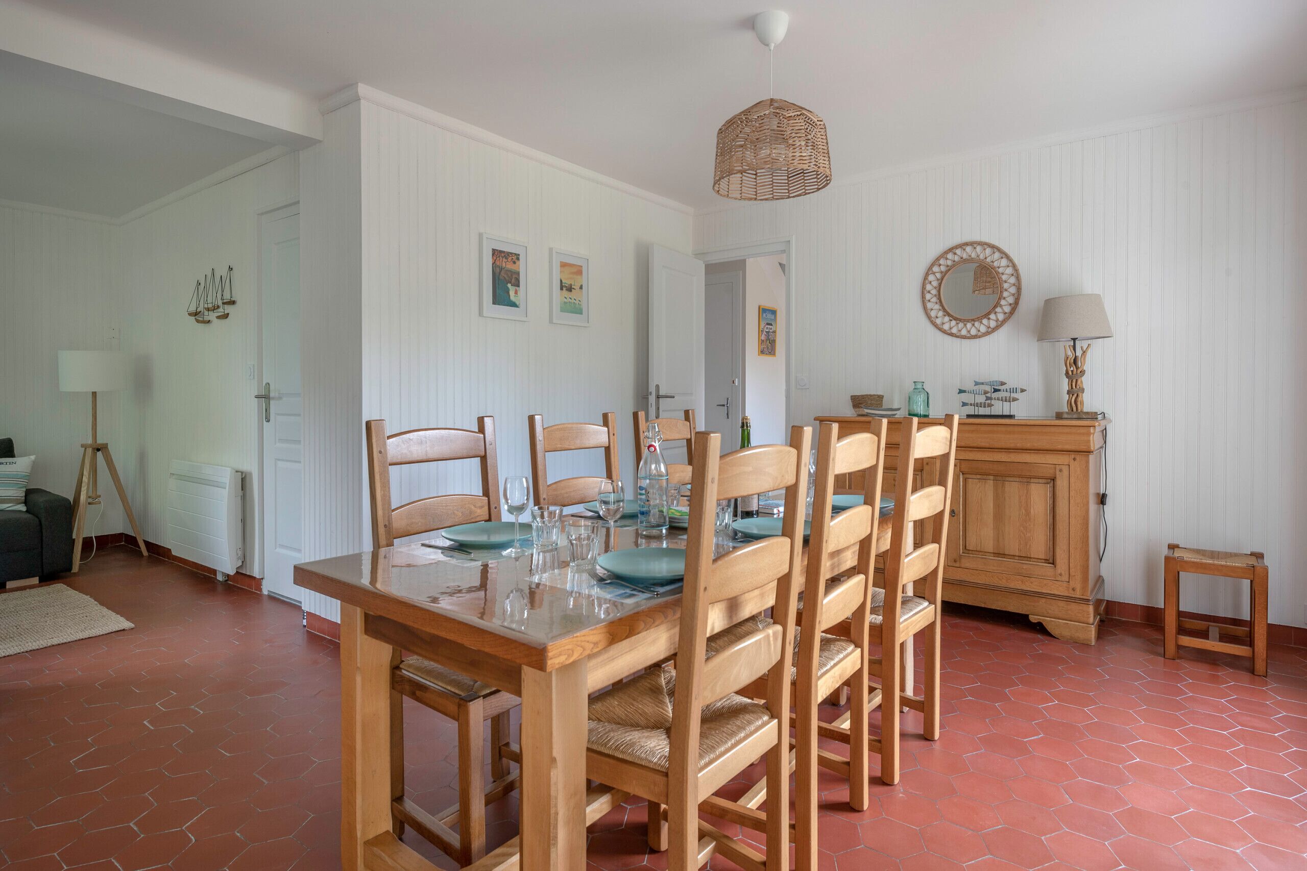 A dining area with a wooden table set for six, surrounded by six wooden chairs. A wooden cabinet and a small side table are visible in the background.