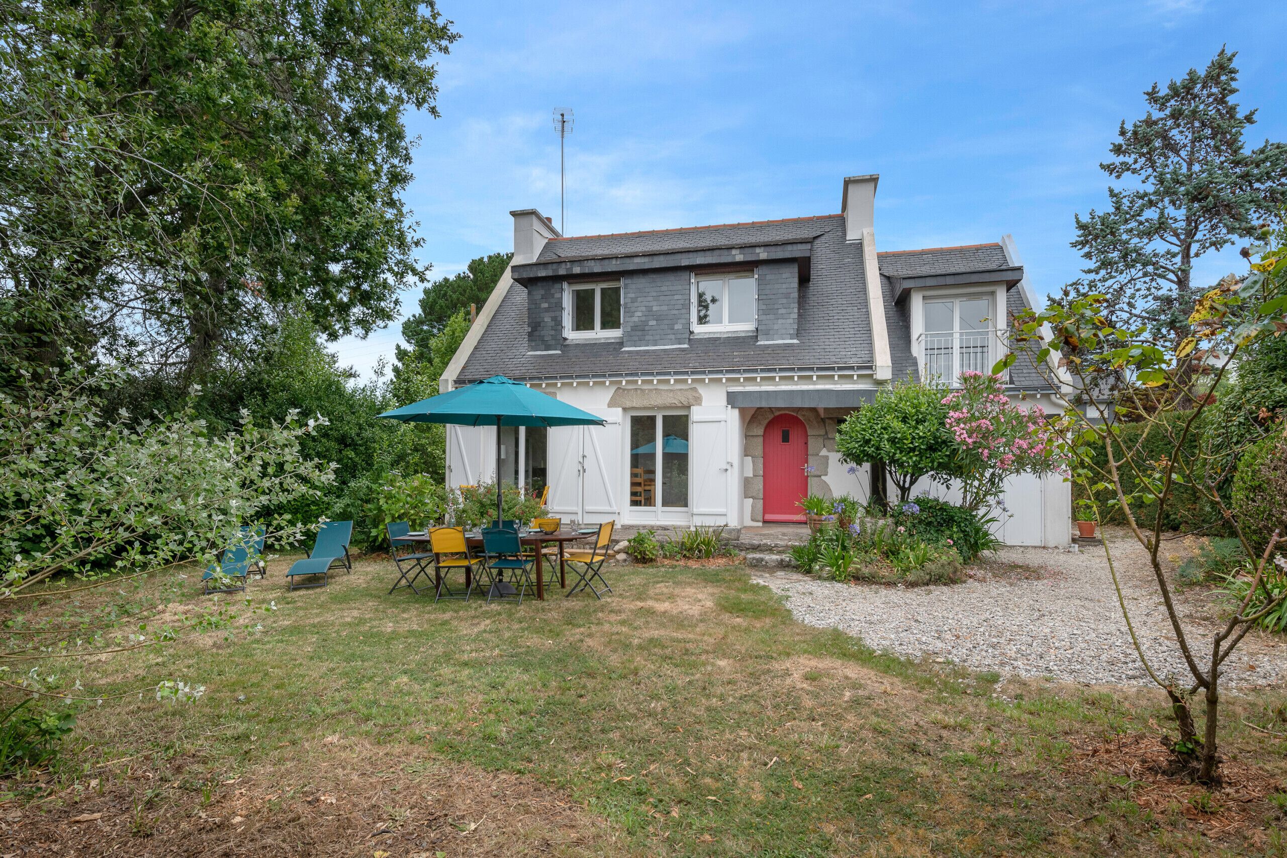 A two-story house with a red door, a covered patio with outdoor furniture, and a garden with various plants and a small tree.