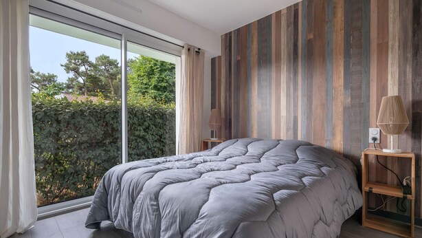 A bedroom with a large bed, a wooden headboard, a nightstand with a lamp, and a view of greenery outside.
