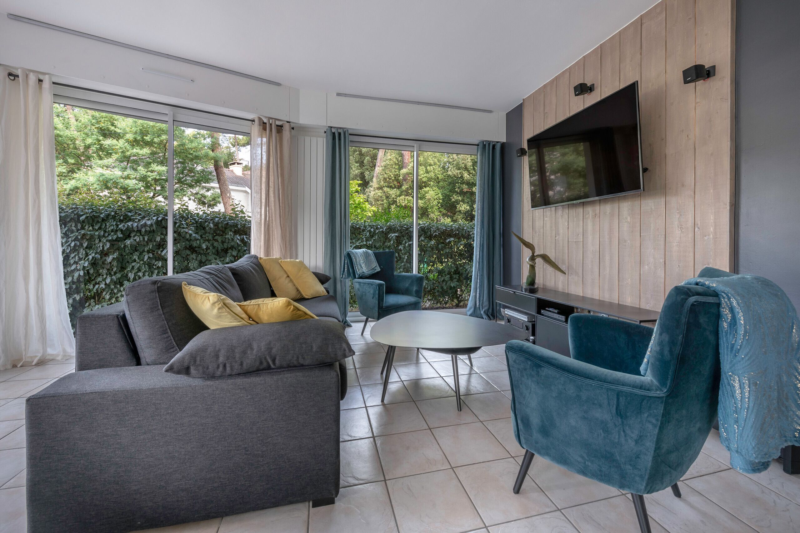 A modern living room with a grey sofa, blue armchairs, and a round coffee table. A flat-screen TV is mounted on the wall.